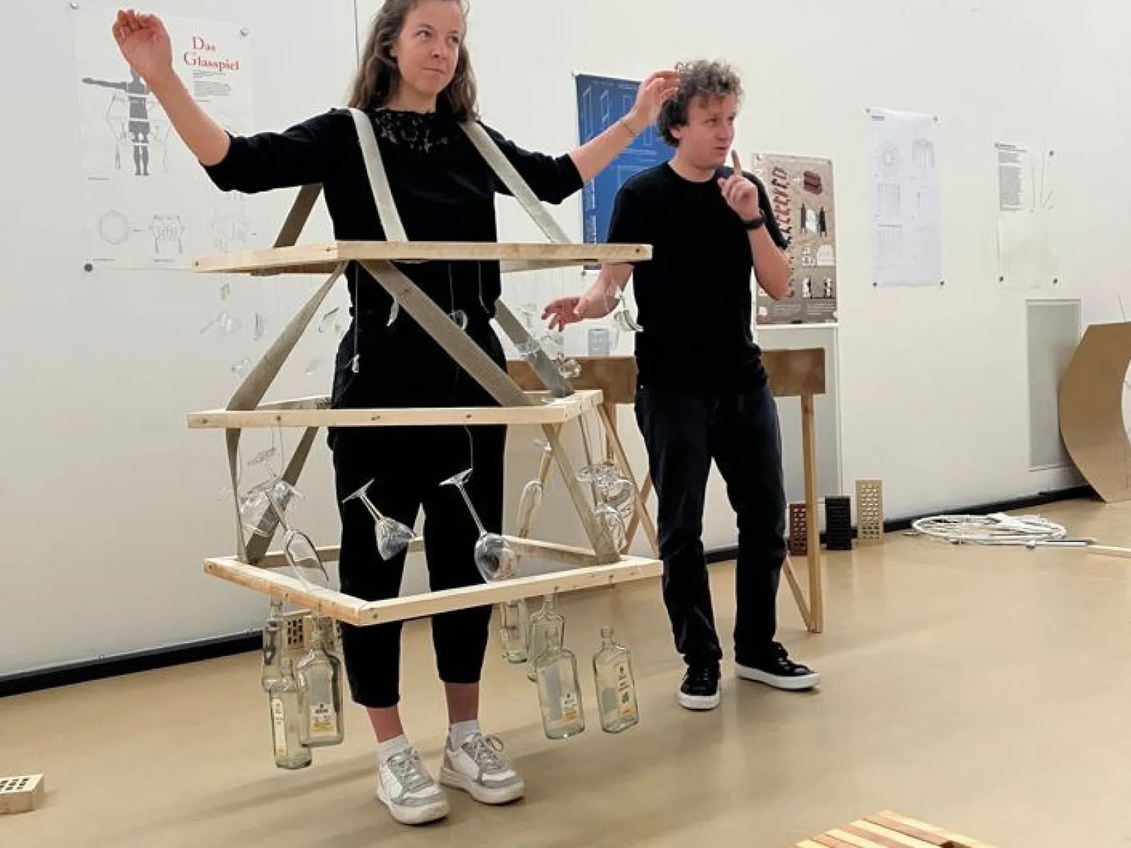 Zwei Studierende präsentieren ein kreatives Designprojekt im Rahmen des Advanced Studio Handwerk an der Universität Liechtenstein. Eine Person trägt eine Konstruktion aus Holzlatten und Glasflaschen. Im Hintergrund sind Plakate und Modelle zu sehen.