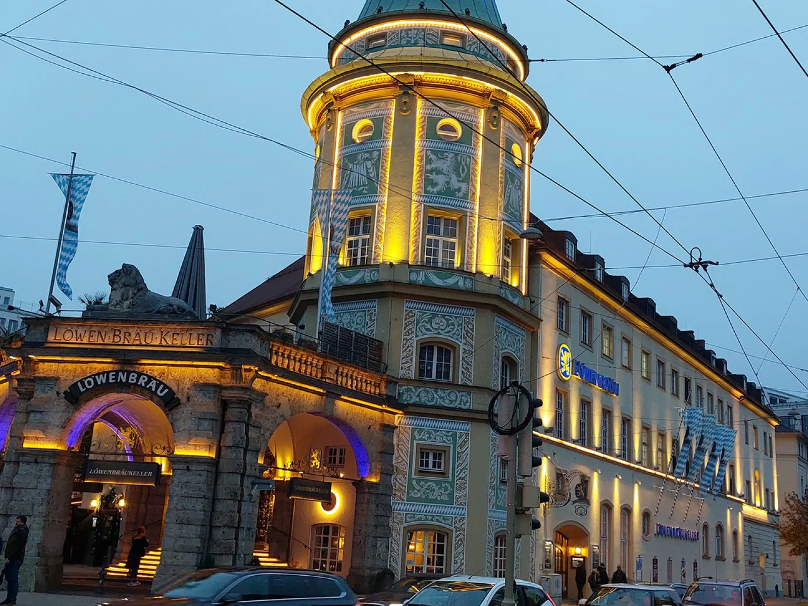 Außenansicht des Löwenbräukellers in München bei Dämmerung, beleuchtet mit warmem Licht, mit vorbeifahrenden Autos und Straßenbahnkabeln im Vordergrund.
