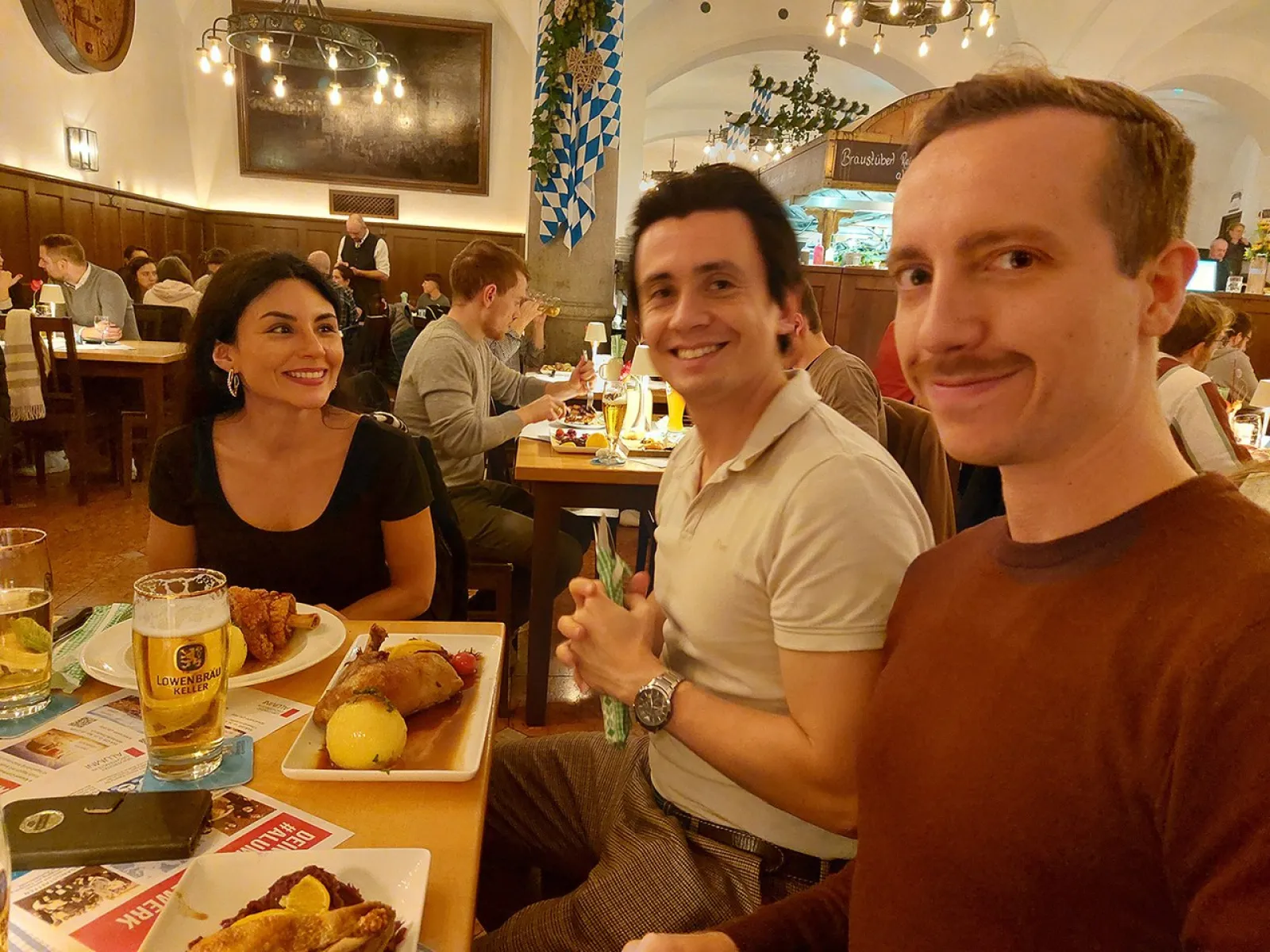 Drei Personen sitzen an einem Tisch in einem Restaurant und lächeln. Auf dem Tisch stehen traditionelle bayerische Speisen und Getränke. Im Hintergrund sind weitere Gäste und eine gemütliche Atmosphäre zu sehen.