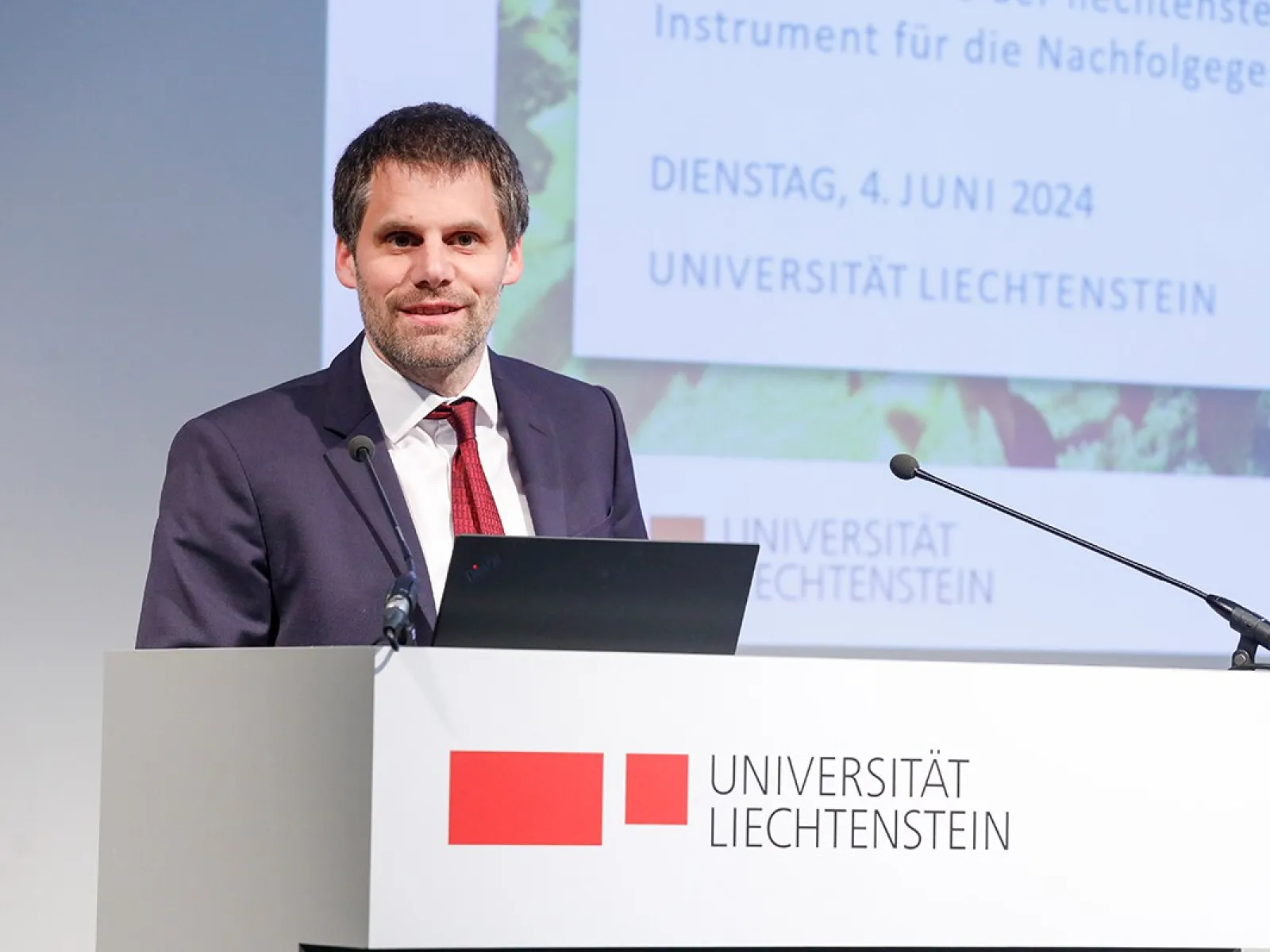 Ein Mann in einem Anzug steht an einem Rednerpult mit dem Logo der Universität Liechtenstein. Im Hintergrund ist eine Präsentationsfolie mit dem Datum 4. Juni 2024 zu sehen.