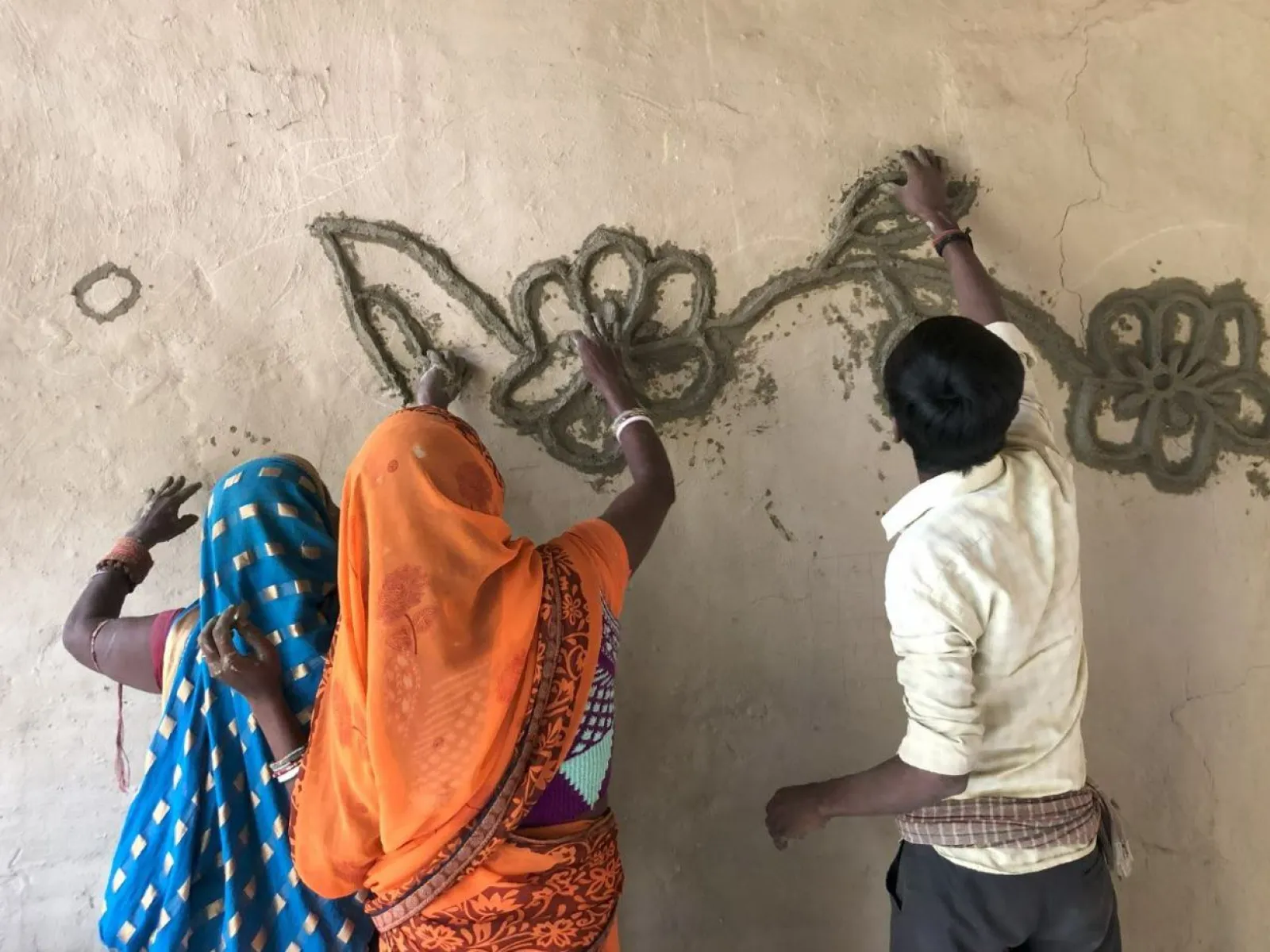 Drei Personen gestalten gemeinsam eine Wand mit traditionellen Mustern aus Lehm. Sie tragen bunte Kleidung und arbeiten konzentriert an einem floralen Design.