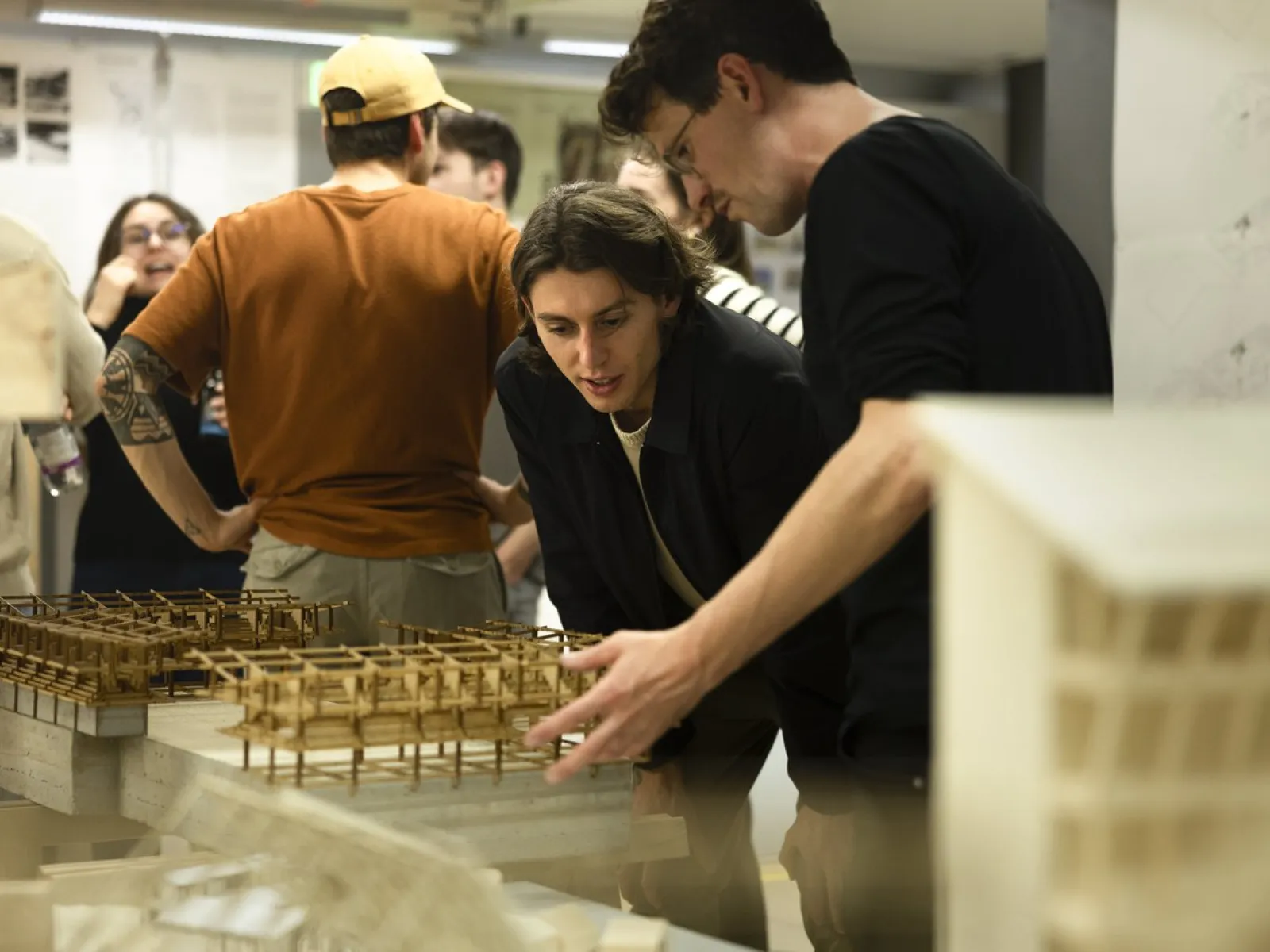 Zwei Personen betrachten aufmerksam Architekturmodelle in einem Ausstellungsraum. Im Hintergrund sind weitere Personen in Gespräche vertieft. Die Szene vermittelt eine Atmosphäre von Interesse und Austausch im Rahmen eines universitären Architektur-Events.