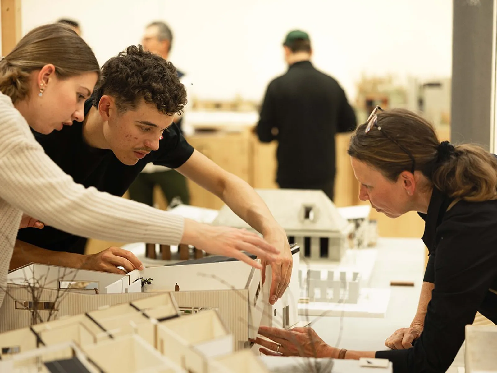 Drei Personen betrachten und diskutieren über Architekturmodelle auf einem Tisch in einem Ausstellungsraum. Im Hintergrund sind weitere Personen und Modelle zu sehen.