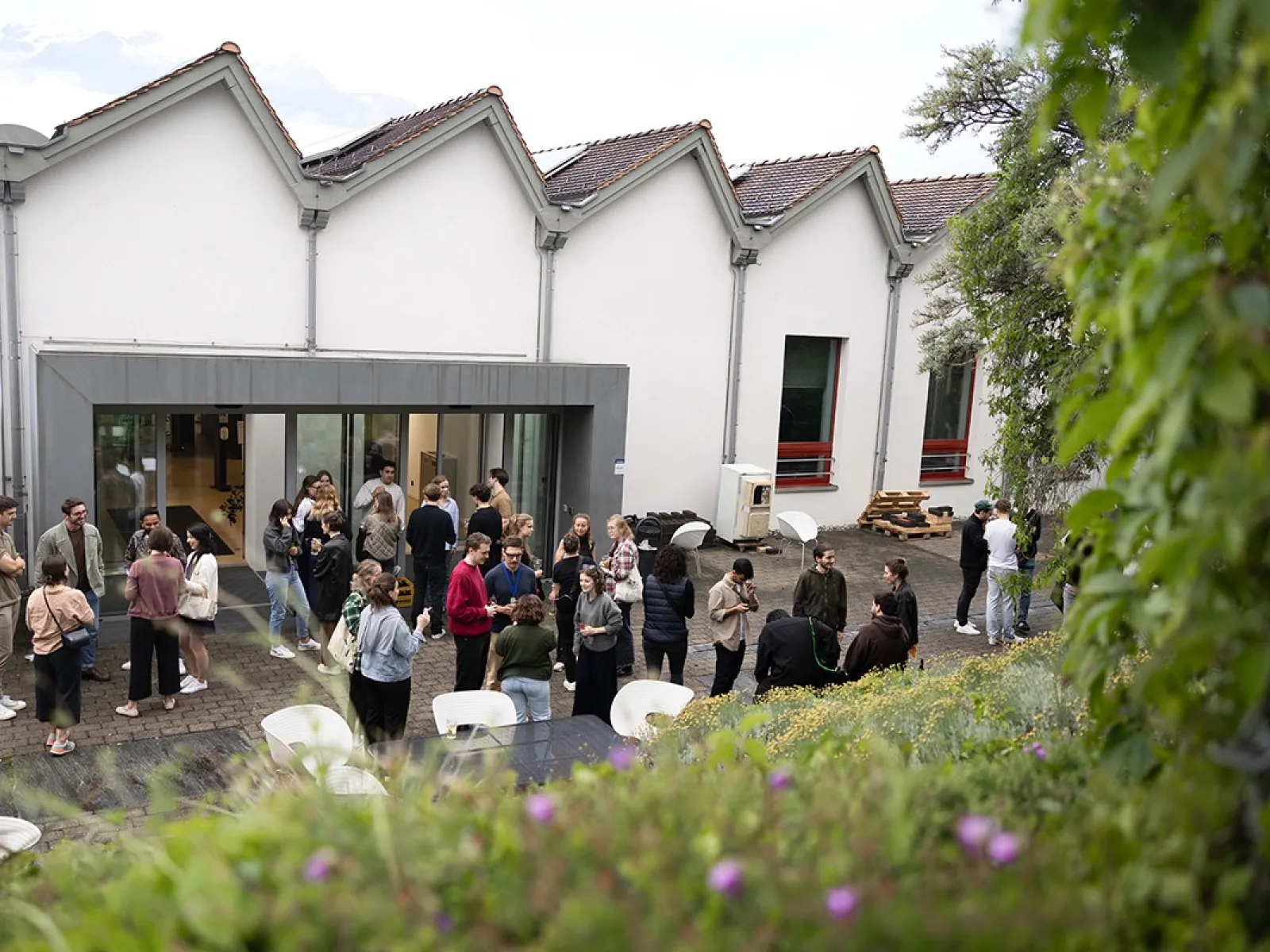 Eine Gruppe von Menschen versammelt sich im Innenhof der Liechtenstein School of Architecture während des Architektur Sommerfests. Im Hintergrund sind moderne Gebäude mit großen Fenstern zu sehen. Die Atmosphäre ist lebhaft und gesellig.