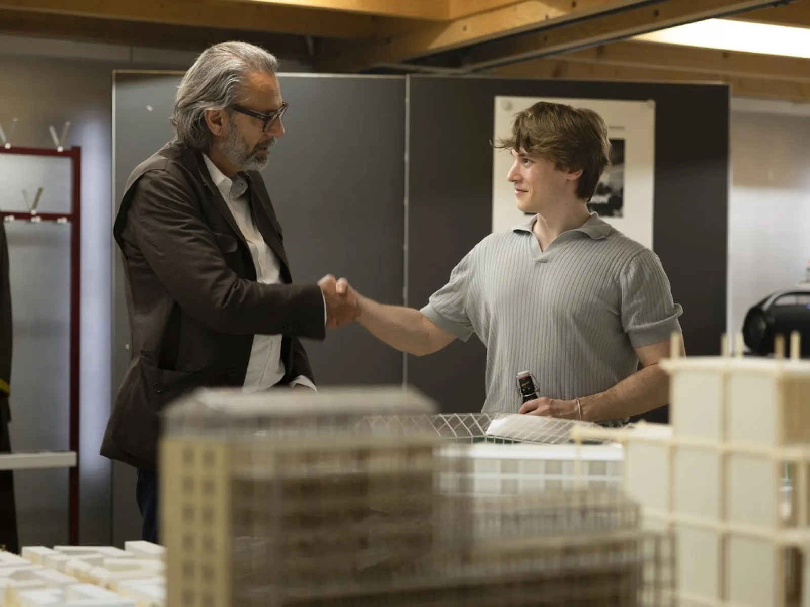 Zwei Personen schütteln sich die Hand in einem Raum mit Architekturmodellen. Eine Person hält ein Getränk. Die Szene vermittelt eine freundliche und professionelle Atmosphäre.