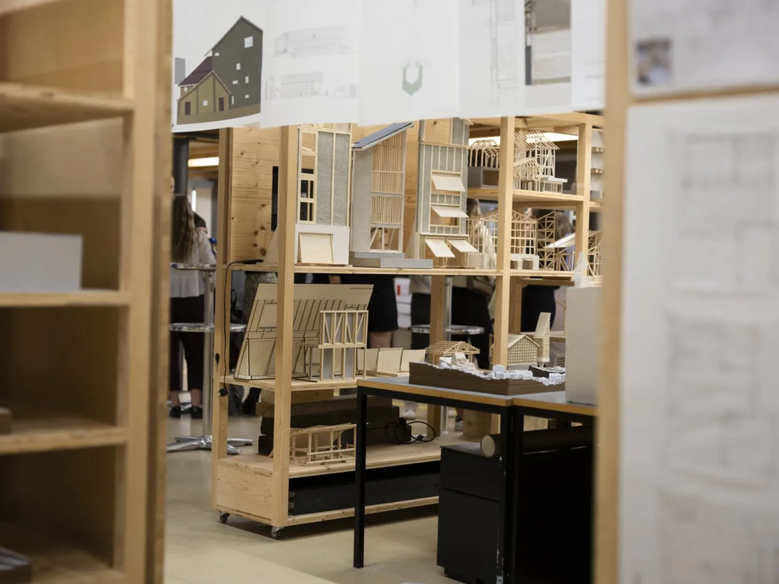 Architekturmodelle und Zeichnungen in einem Ausstellungsraum der Universität Liechtenstein. Holzregale präsentieren detaillierte Miniaturgebäude und Baupläne. Im Hintergrund sind Personen unscharf zu erkennen.