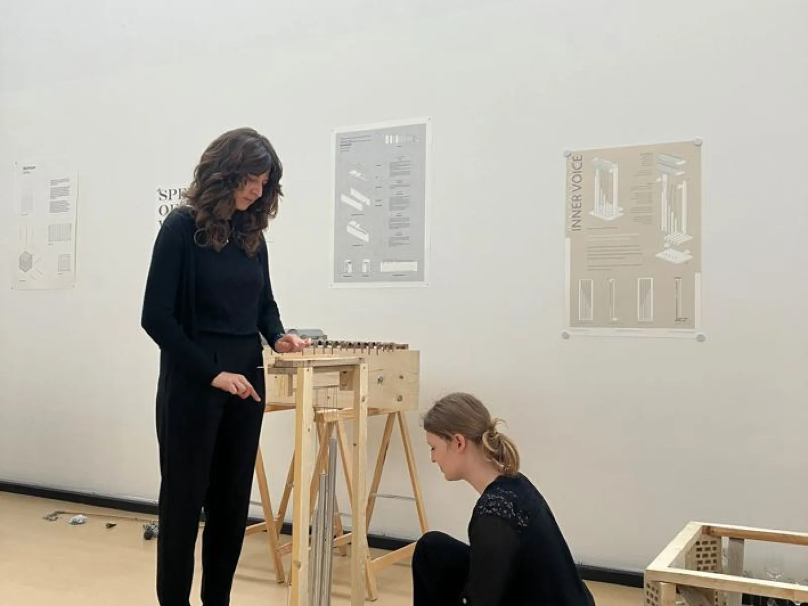 Zwei Studierende arbeiten in einem hellen Raum an einem architektonischen Modell. Im Hintergrund sind Poster mit Designplänen an der Wand zu sehen.