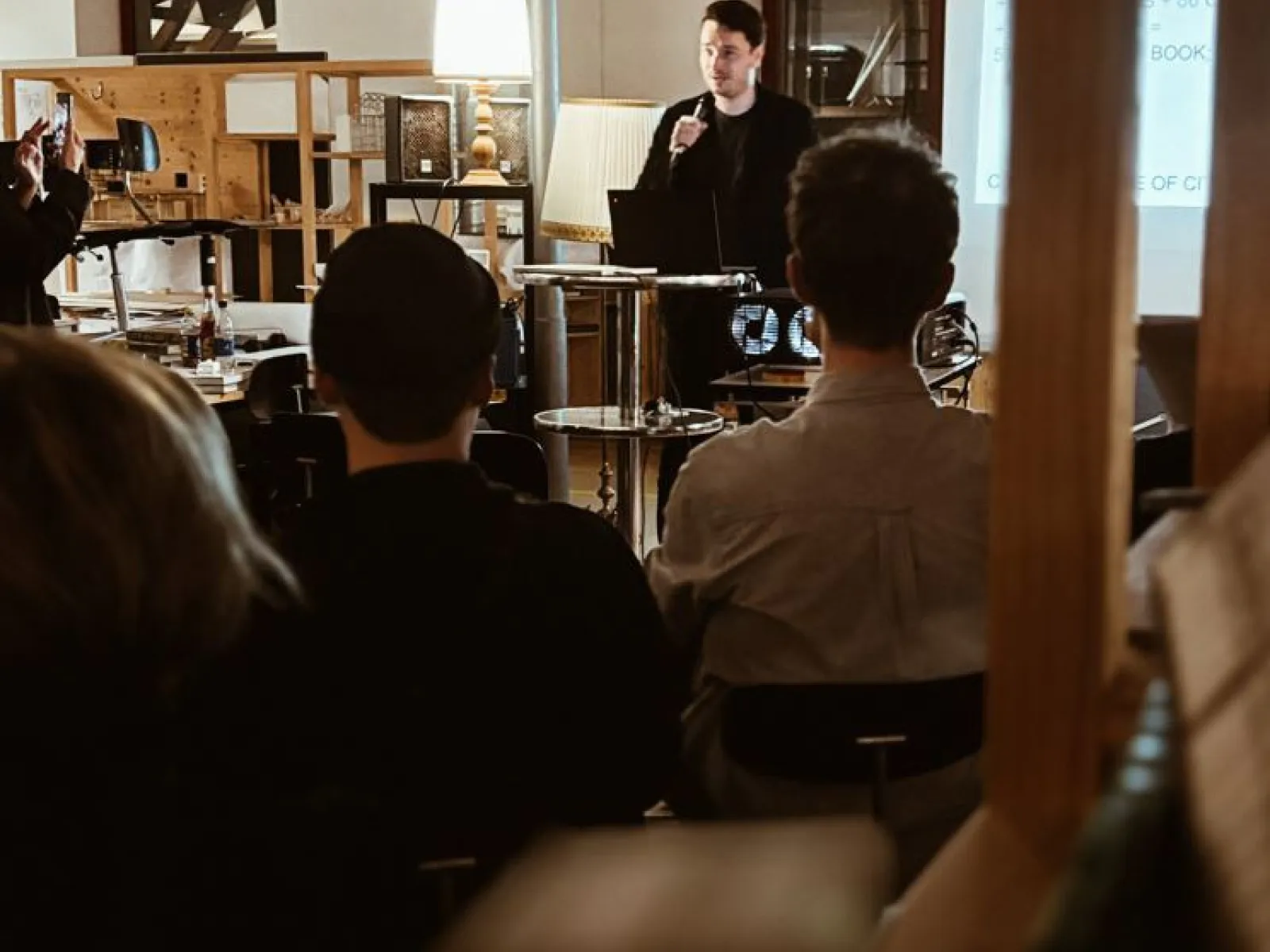 Ein Vortragender spricht vor einer Gruppe von Studierenden in einem Raum mit Präsentationstechnik. Im Hintergrund sind Regale und eine Lampe sichtbar. Die Atmosphäre wirkt konzentriert und lernorientiert.