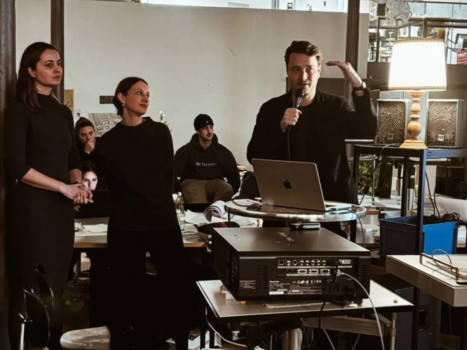 Eine Gruppe von Personen steht in einem Raum mit moderner Einrichtung. Eine Person hält ein Mikrofon und spricht, während andere zuhören. Im Hintergrund sind Tische, Stühle und technische Geräte zu sehen. Die Szene wirkt wie eine Präsentation oder ein Workshop in einem akademischen Umfeld.