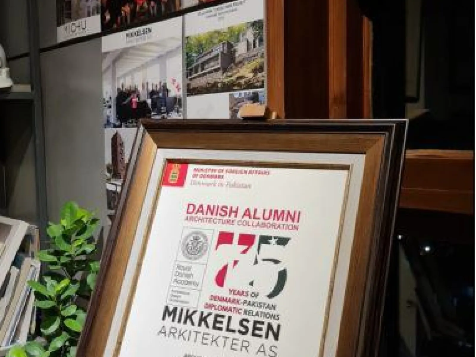 Ein gerahmtes Zertifikat zur Anerkennung der Zusammenarbeit im Bereich Architektur zwischen Dänemark und Pakistan, ausgestellt an Fasih D. Khan, steht auf einem Regal. Im Hintergrund sind Architekturprojekte und Gruppenfotos zu sehen.
