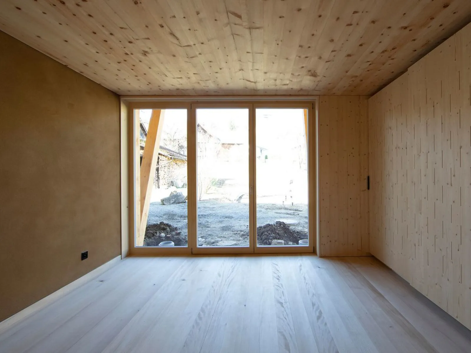 Innenansicht eines minimalistischen Raumes mit Holzverkleidung und großem Fenster, das den Blick auf eine ländliche Umgebung freigibt. Der Raum ist lichtdurchflutet und zeigt eine moderne, nachhaltige Architektur.