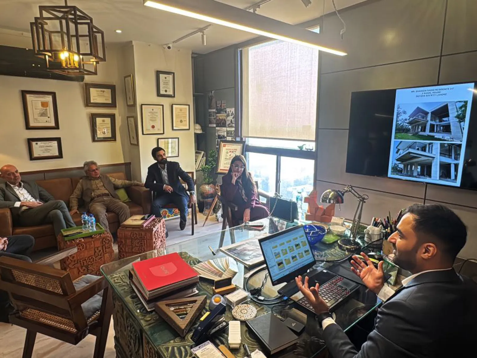 Ein Büro mit mehreren Personen, die in einem Meeting sitzen. Eine Person sitzt an einem Schreibtisch mit einem Laptop, während andere auf Stühlen und einem Sofa Platz genommen haben. An der Wand hängen Zertifikate und ein Bildschirm zeigt architektonische Entwürfe.