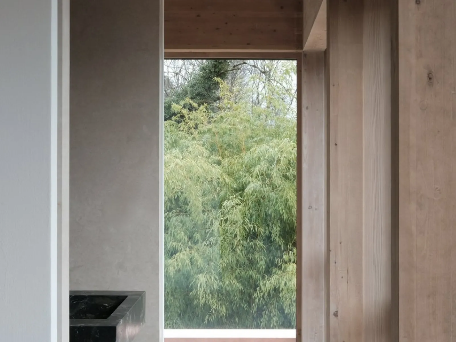 Innenraum mit Holzverkleidung und großem Fenster, das den Blick auf grüne Bäume freigibt. Moderne Architektur mit Fokus auf natürliche Materialien und Licht.