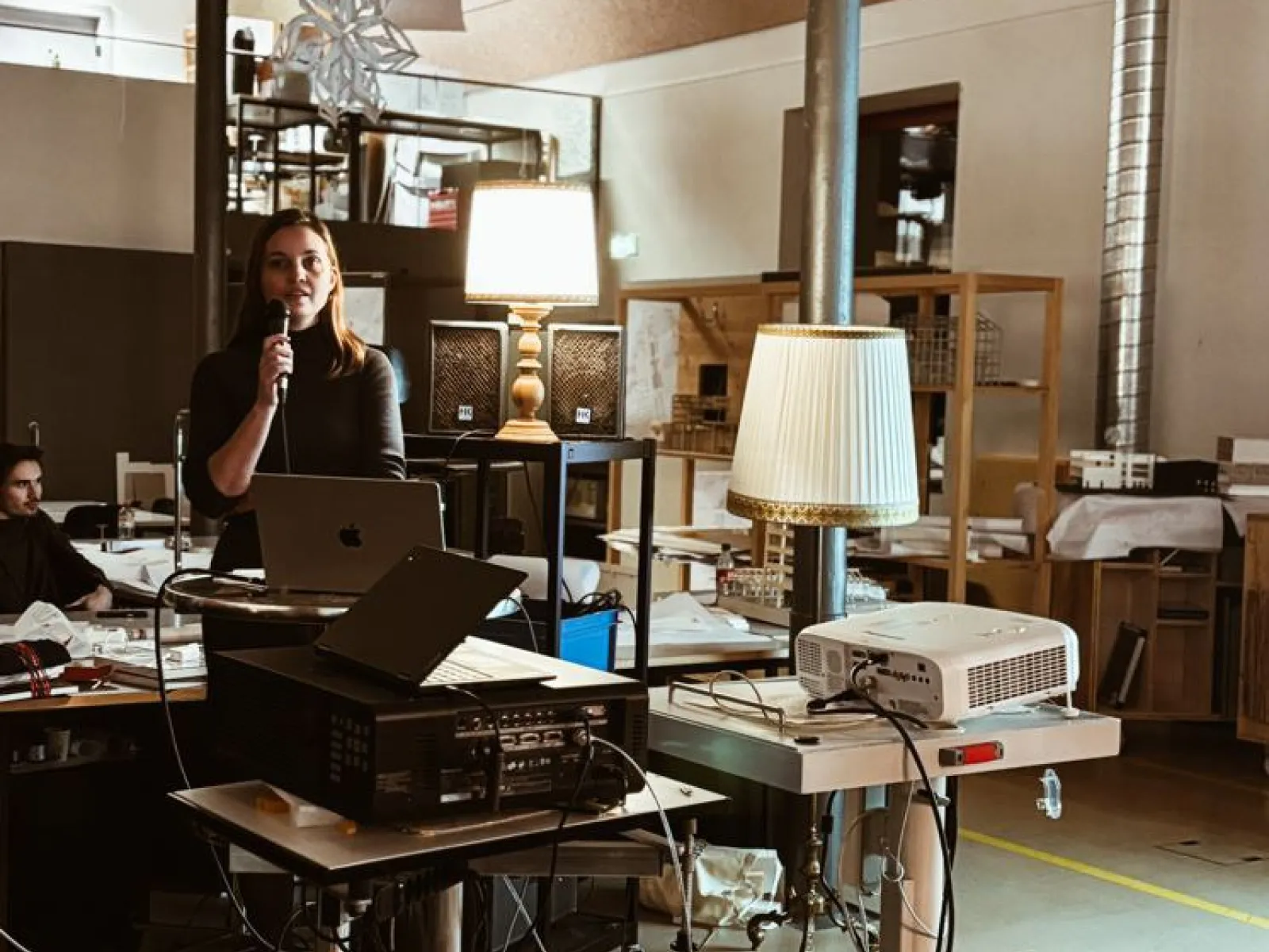 Eine Person hält eine Präsentation in einem modernen Raum mit technischen Geräten und Lampen. Im Hintergrund sind Regale und Arbeitsmaterialien zu sehen.