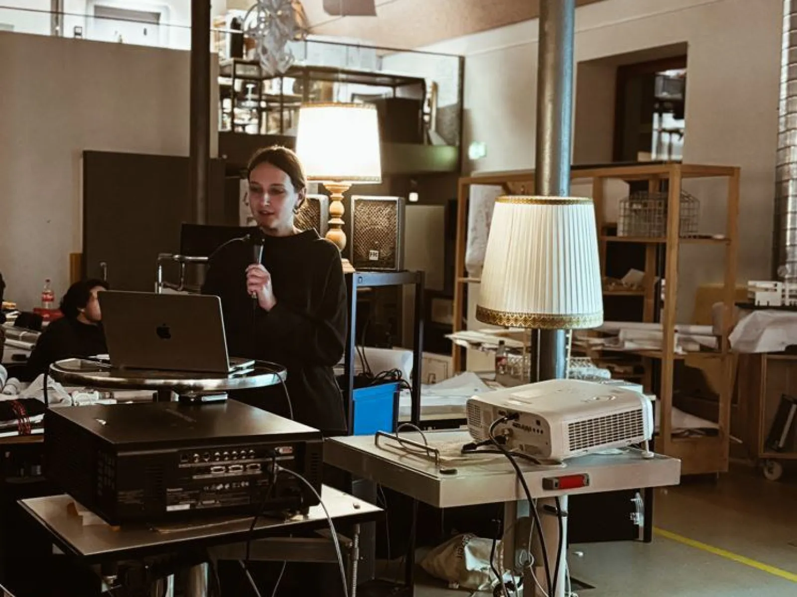 Eine Person hält eine Präsentation in einem modernen Raum mit technischer Ausstattung. Im Vordergrund stehen ein Laptop und ein Projektor auf einem Tisch. Der Raum ist gut beleuchtet und im Hintergrund sind Regale und weitere Personen zu sehen.