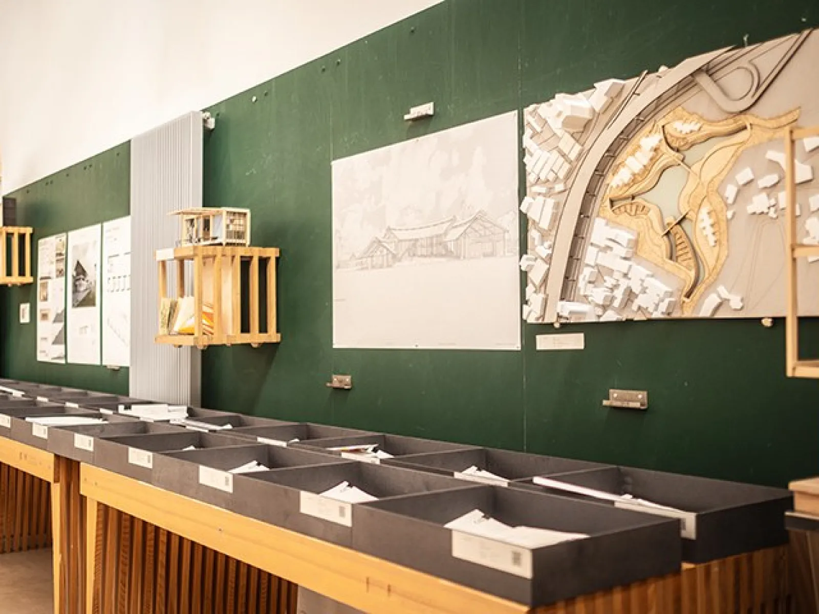 Architekturmodelle und Zeichnungen an einer grünen Pinnwand in einem Raum der Liechtenstein School of Architecture. Im Vordergrund sind Ablagefächer mit Dokumenten sichtbar.