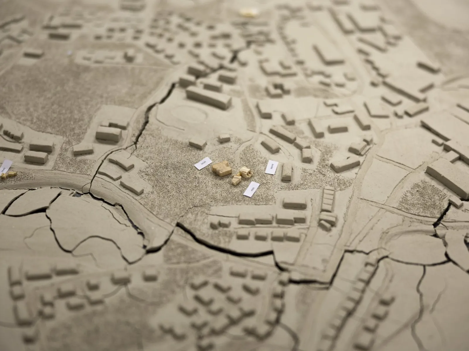 Ein detailliertes Architekturmodell zeigt eine Stadtlandschaft mit Gebäuden und Straßen. Im Zentrum sind mehrere kleine, beschriftete Modellelemente platziert, die möglicherweise auf ein spezifisches Bauprojekt hinweisen. Das Modell bietet eine Vogelperspektive auf die städtische Umgebung und dient als visuelles Hilfsmittel für architektonische Planungen und Präsentationen.