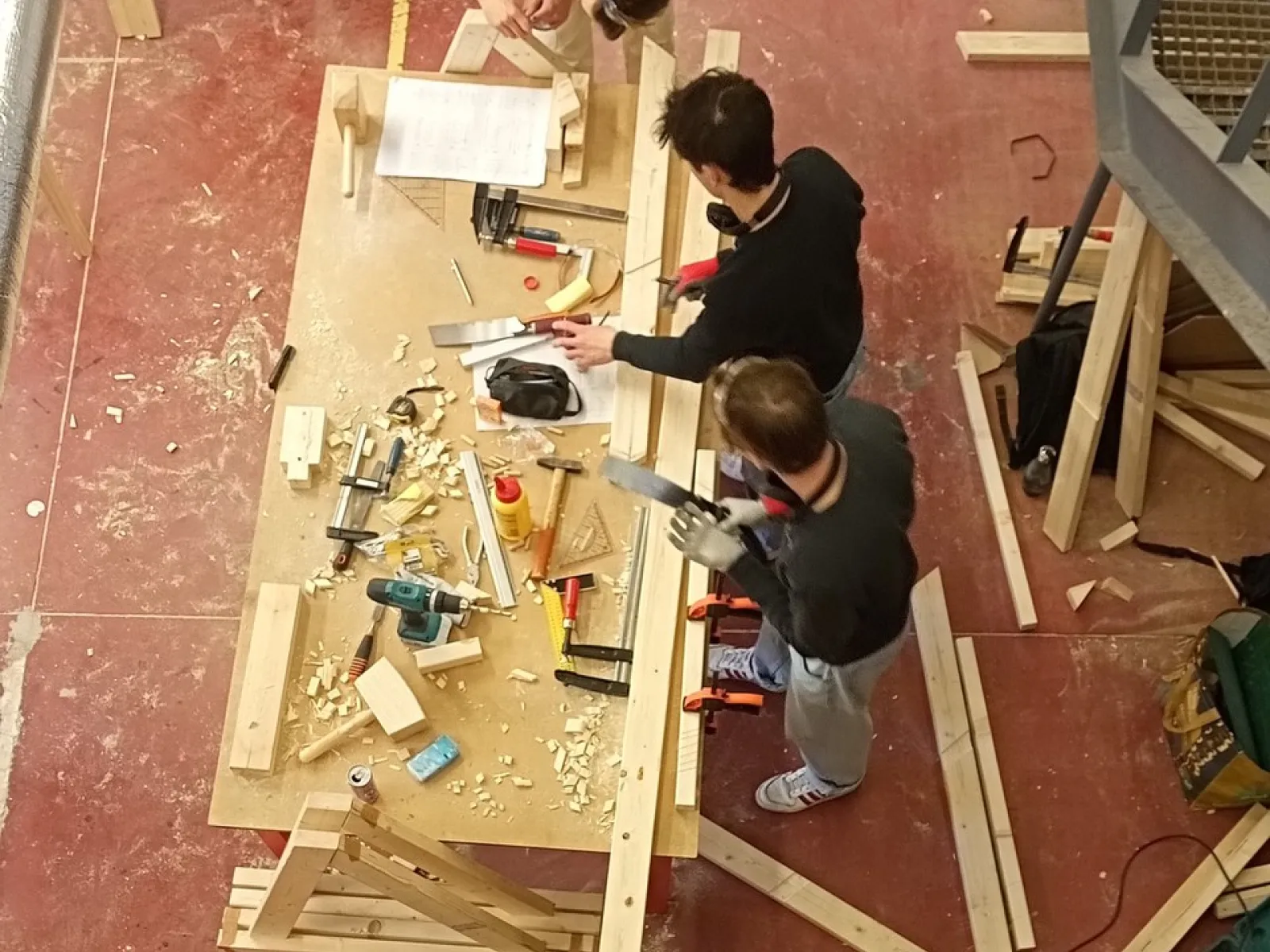 Vier Studierende arbeiten gemeinsam in einer Werkstatt an einem Holztisch. Sie sind von oben zu sehen und verwenden verschiedene Werkzeuge, um ein Möbelstück zu bauen. Holzstücke und Werkzeuge liegen auf dem Tisch verteilt.