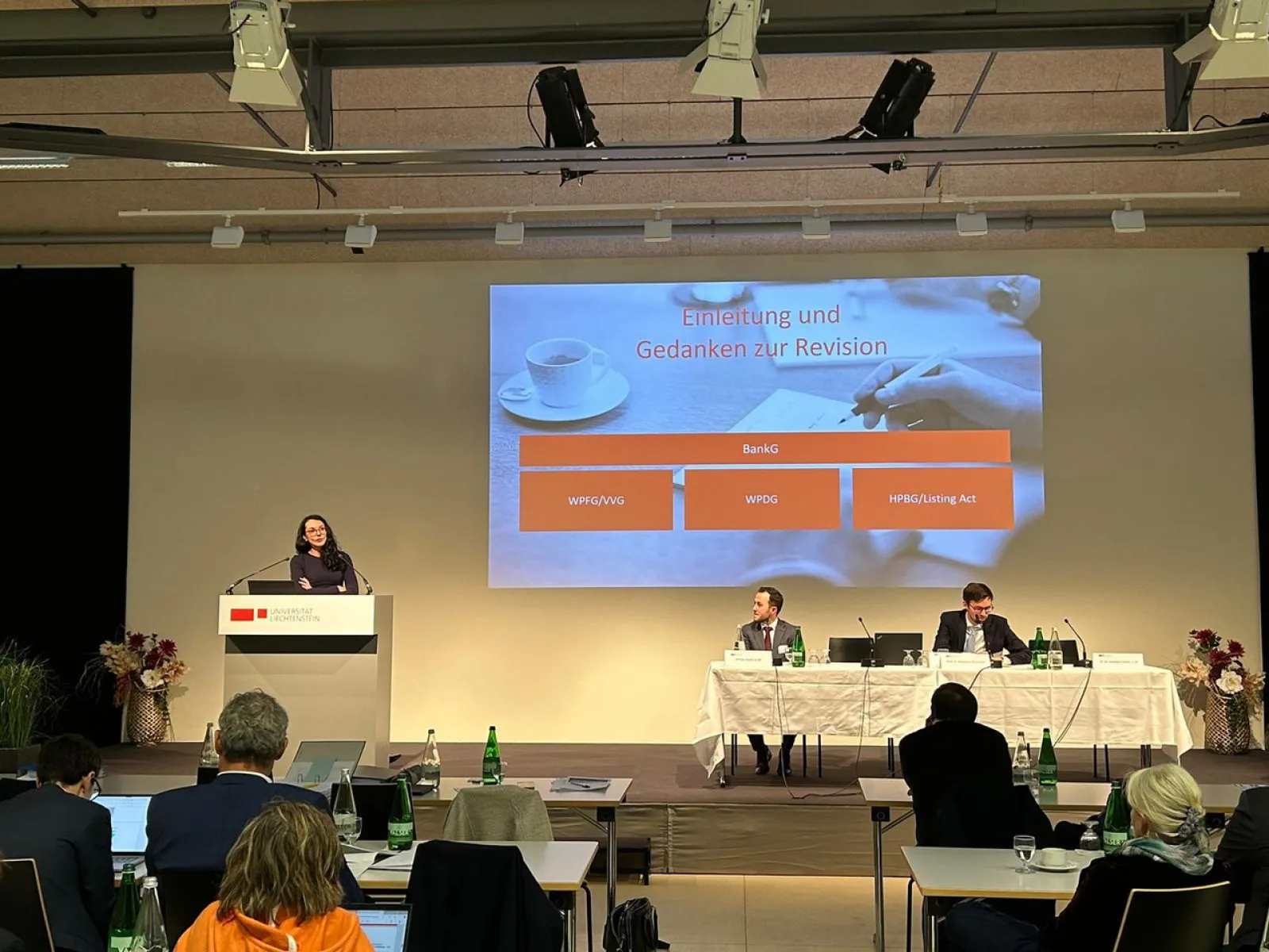 Eine Person hält einen Vortrag auf einer Bühne beim Liechtensteinischen Bankrechtsforum an der Universität Liechtenstein. Im Hintergrund ist eine Präsentationsfolie mit dem Titel "Einleitung und Gedanken zur Revision" zu sehen. Zwei weitere Personen sitzen an einem Tisch auf der Bühne. Im Publikum sind mehrere Zuhörer.
