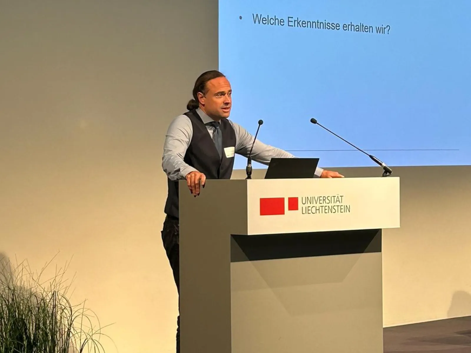 Ein Redner steht an einem Podium mit dem Logo der Universität Liechtenstein. Im Hintergrund ist eine Präsentationsfolie mit dem Titel "Reflexion" und den Fragen "Wo stehen wir?", "Was betrachten wir?" und "Welche Erkenntnisse erhalten wir?" zu sehen. Links im Bild sind Pflanzen dekorativ platziert.