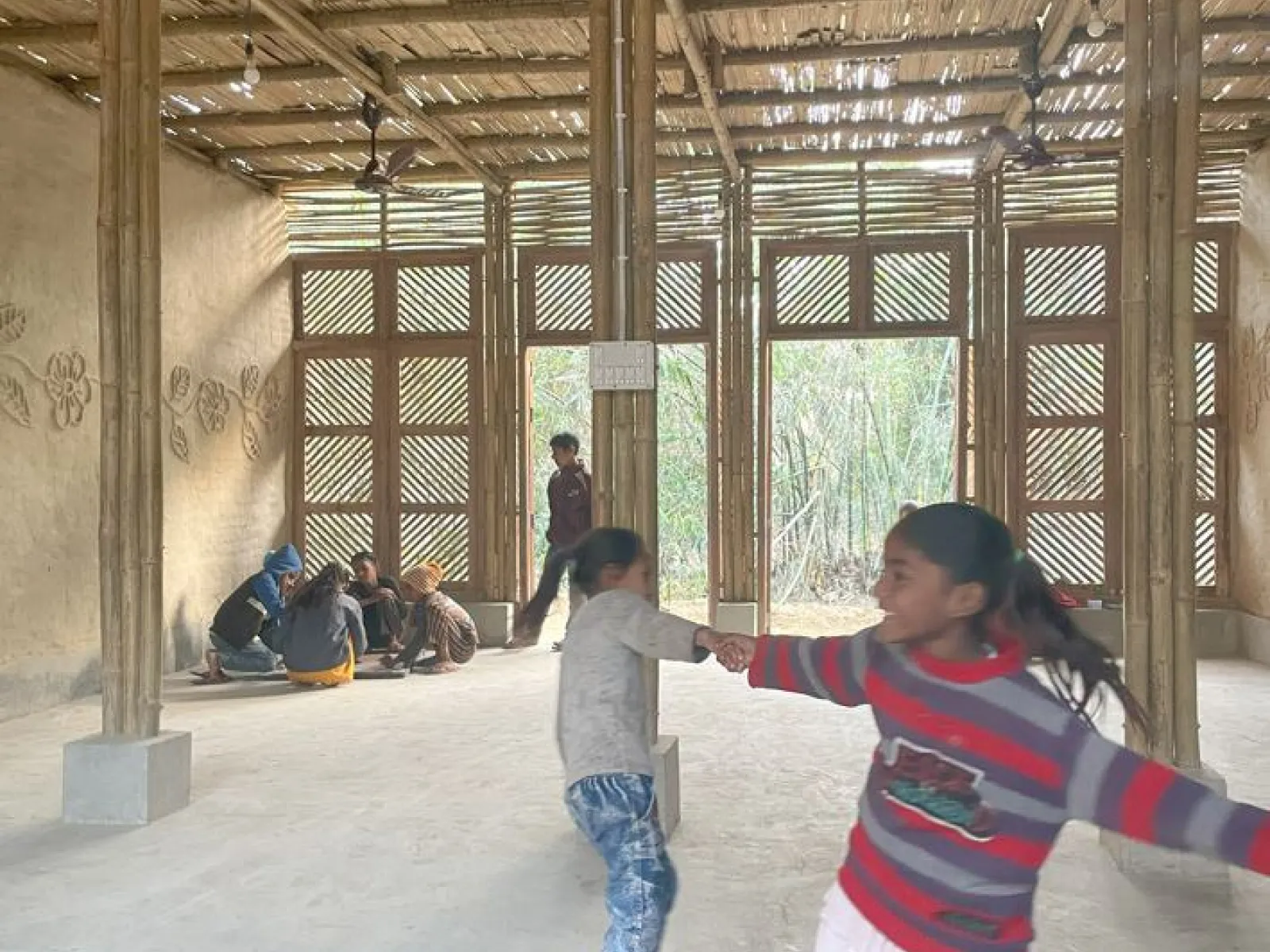 Kinder spielen und tanzen in einem neu gebauten Schulgebäude aus Bambus in Bihar, Indien. Im Hintergrund sitzen weitere Kinder auf dem Boden. Das Gebäude hat eine natürliche, lichtdurchflutete Atmosphäre.