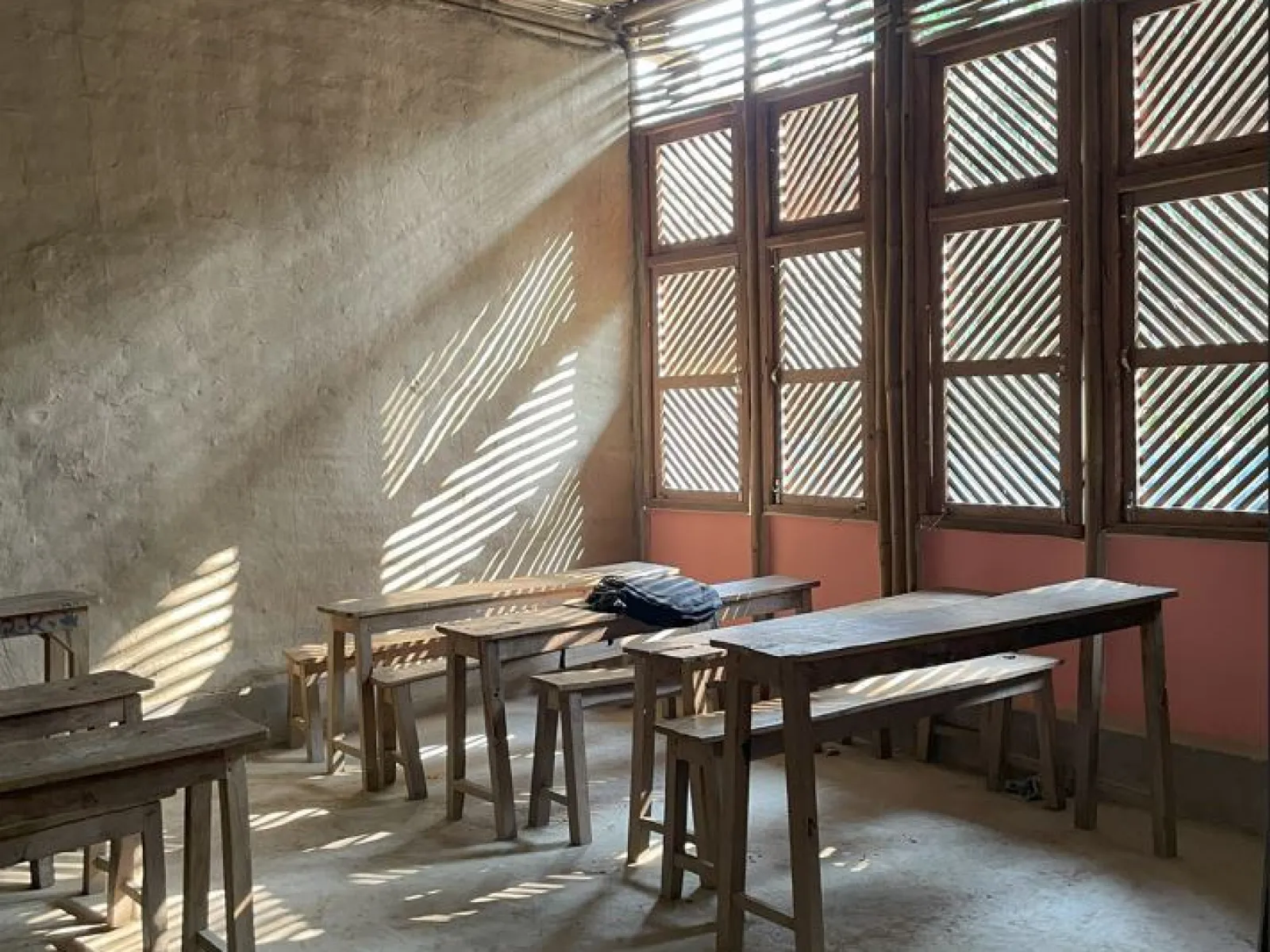 Ein einfacher Klassenraum mit Holztischen und -bänken, durchflutet von Sonnenlicht, das durch Fenster mit Holzlamellen fällt.