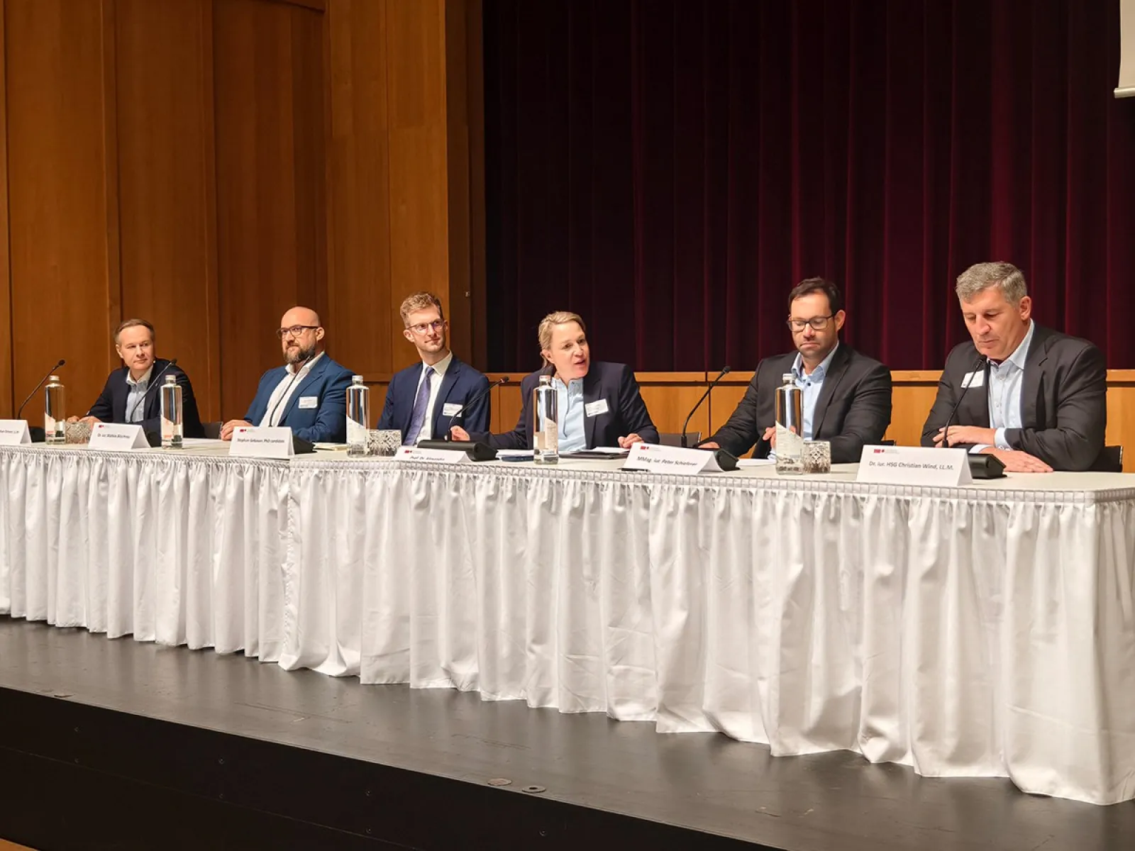Eine Gruppe von sechs Personen sitzt an einem langen Tisch auf einer Bühne. Sie tragen formelle Kleidung und haben Namensschilder vor sich. Im Hintergrund sind Vorhänge zu sehen. Der Tisch ist mit einem weißen Tuch bedeckt und es stehen Wasserflaschen darauf.