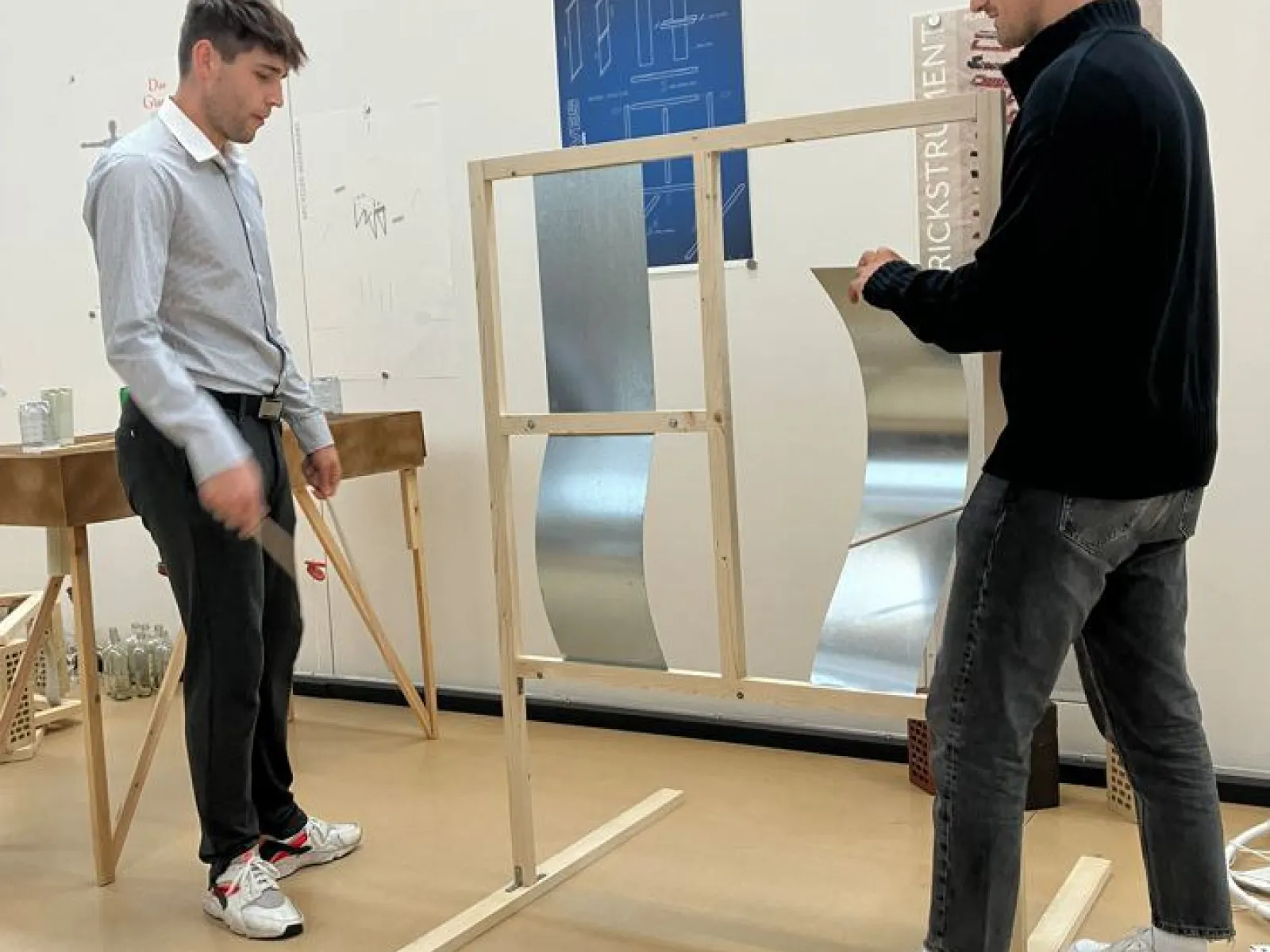 Zwei Studierende arbeiten in einem Atelier an einem Holzrahmen mit Metallteilen. Im Hintergrund sind Pläne und Modelle zu sehen.