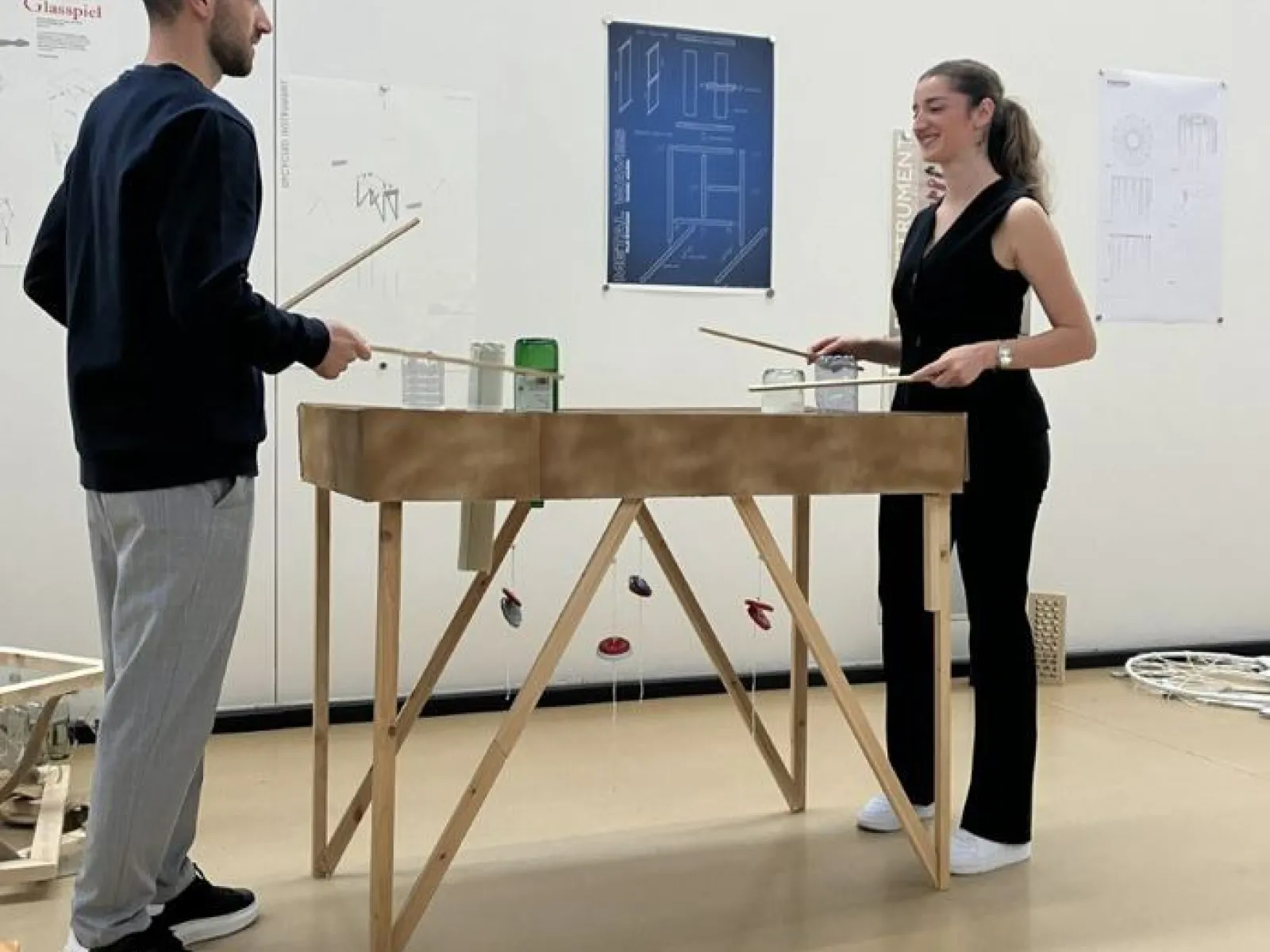 Zwei Personen stehen an einem hölzernen Tischmodell und diskutieren. Im Hintergrund sind Architekturzeichnungen an der Wand zu sehen. Die Szene spielt in einem Designstudio der Universität Liechtenstein.