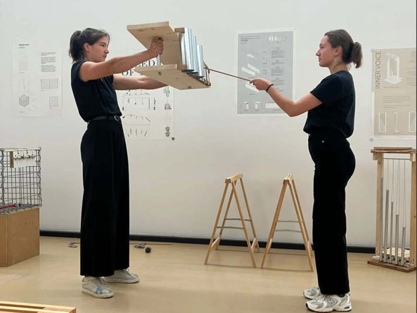 Zwei Studierende in einem Designstudio präsentieren ein Modell. Sie stehen sich gegenüber und halten gemeinsam eine Konstruktion aus Holz und Metall. Im Hintergrund sind Plakate mit architektonischen Entwürfen zu sehen.