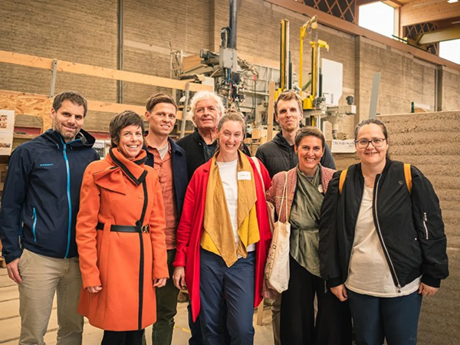 Eine Gruppe von acht Personen steht in einer Werkhalle vor einer Maschine und einem Stampflehm-Element. Sie lächeln in die Kamera.