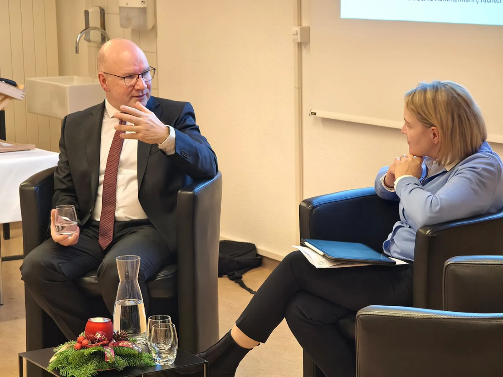 Zwei Personen sitzen in Sesseln und führen ein Gespräch. Der Mann links trägt einen Anzug und hält ein Glas, während er gestikuliert. Die Frau rechts hält ein Notizbuch. Im Vordergrund steht ein Tisch mit einer Karaffe und einem weihnachtlichen Gesteck.