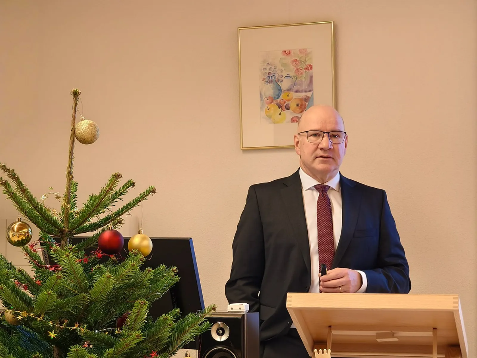Ein Mann in Anzug und Krawatte steht an einem Rednerpult in einem festlich dekorierten Raum mit einem Weihnachtsbaum. Im Hintergrund hängt ein Bild an der Wand.