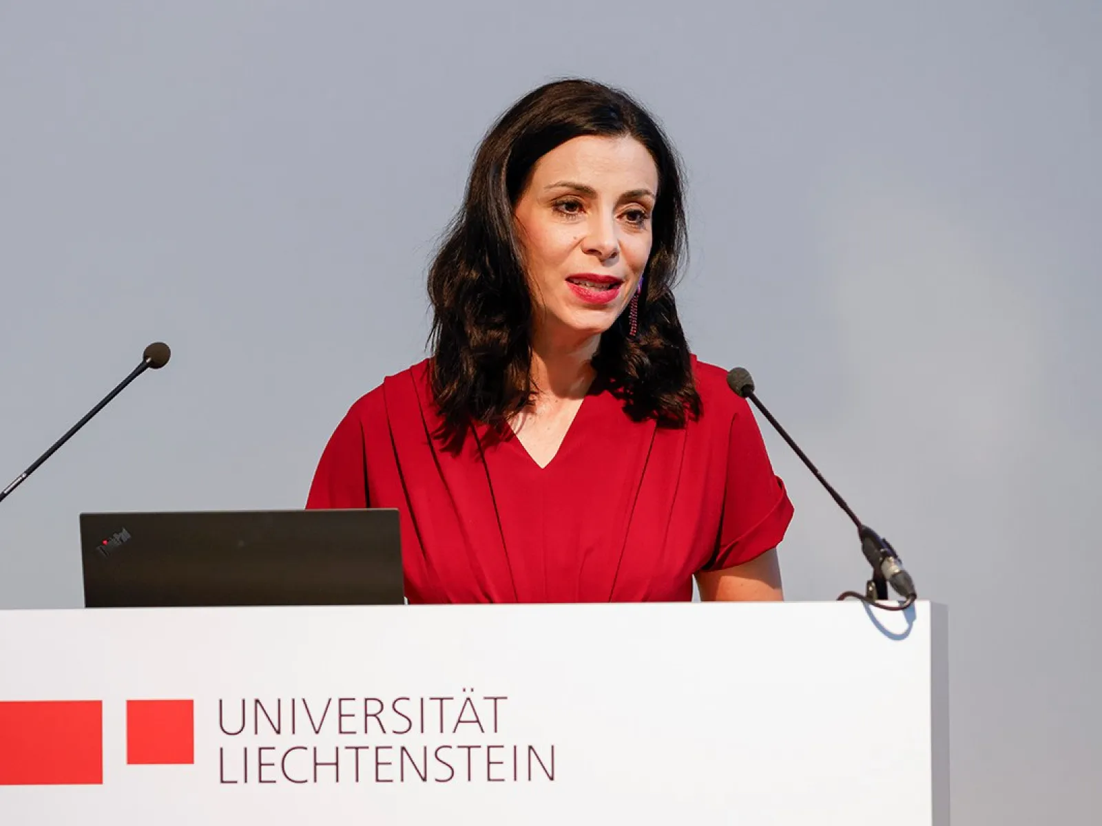 Eine Frau in rotem Kleid spricht an einem Rednerpult mit dem Logo der Universität Liechtenstein. Sie hält eine Rede vor einem Mikrofon.