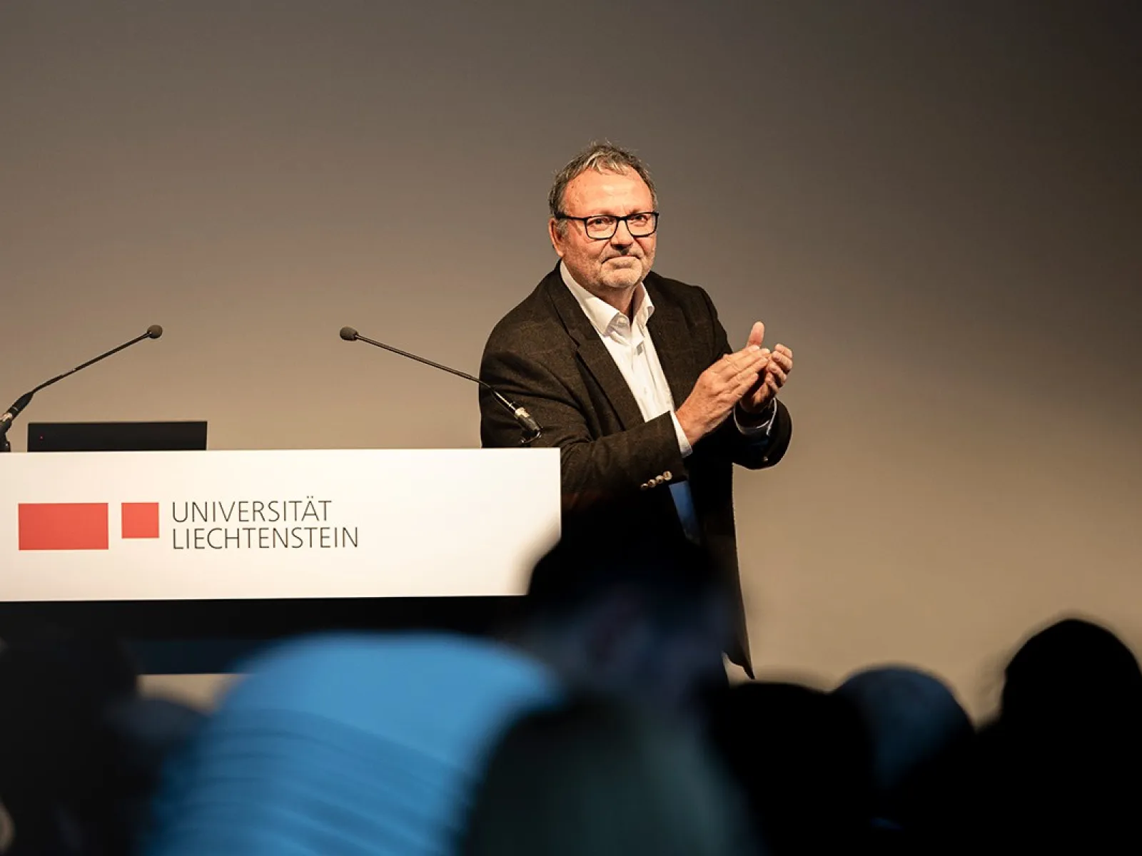 Ein Mann steht an einem Rednerpult der Universität Liechtenstein und applaudiert. Im Hintergrund sind Mikrofone sichtbar.