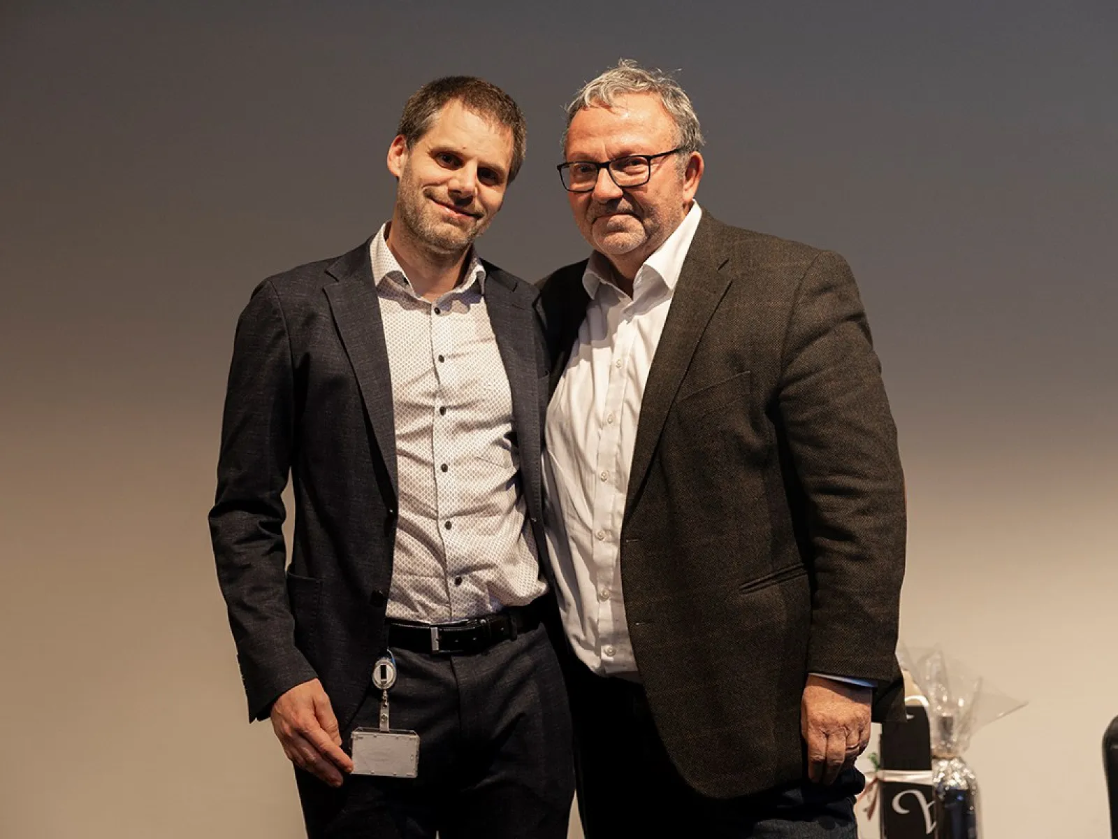Zwei Männer stehen nebeneinander und lächeln in die Kamera. Sie tragen Anzüge und befinden sich in einem Innenraum mit neutralem Hintergrund.