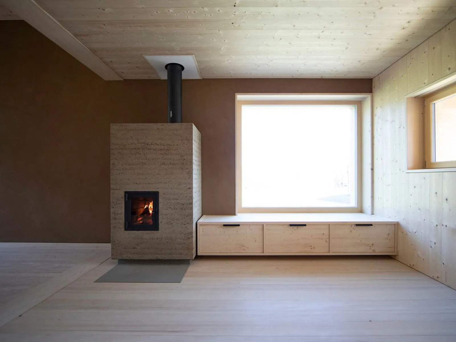Ein modernes, minimalistisches Wohnzimmer mit einem Holzofen, einer großen Fensterbank und Holzwänden, gestaltet von EMM Architekten.