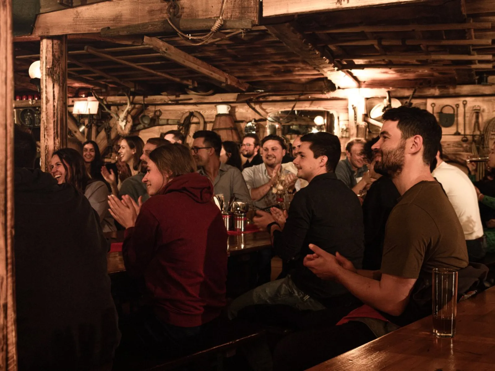 Eine Gruppe von Menschen sitzt in einem rustikalen Raum und applaudiert während einer Veranstaltung. Die Atmosphäre ist gemütlich und gesellig.