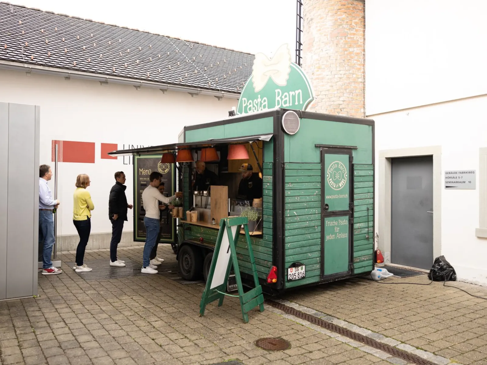 Ein grüner Foodtruck mit der Aufschrift "Pasta Barn" steht auf einem gepflasterten Platz. Mehrere Personen stehen in einer Schlange vor dem Truck und warten auf ihre Bestellung. Im Hintergrund sind Gebäude der Universität Liechtenstein zu sehen.
