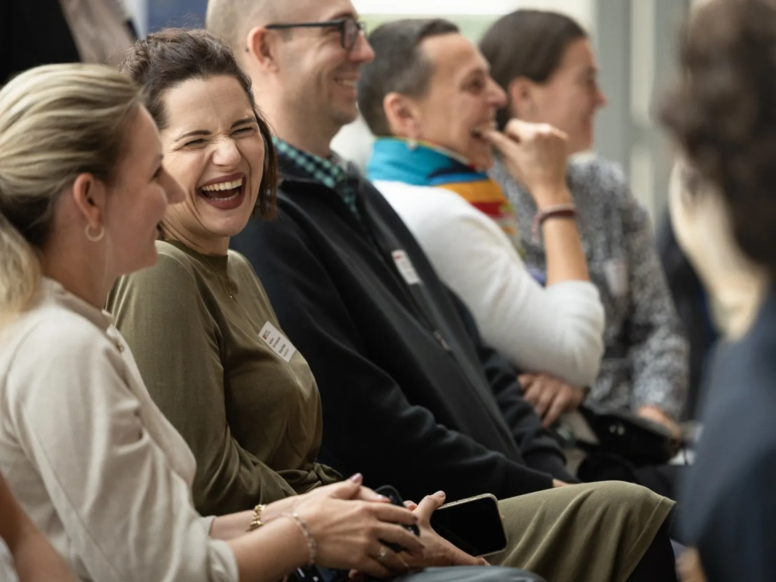 Eine Gruppe von Menschen sitzt lachend in einem Veranstaltungsraum. Sie wirken entspannt und fröhlich, während sie einem Vortrag oder einer Präsentation folgen.