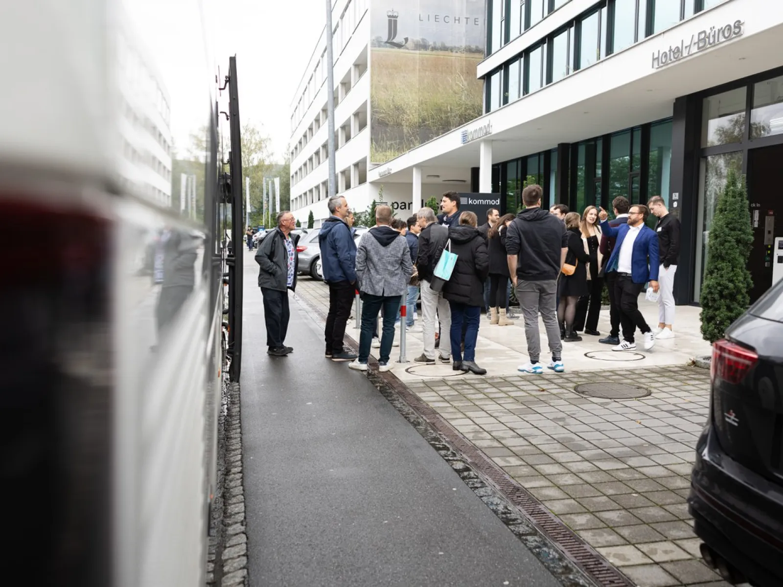 Gruppe von Menschen steht vor einem modernen Gebäude auf dem Campus der Universität Liechtenstein, möglicherweise während einer Veranstaltung. Im Vordergrund sind geparkte Autos zu sehen.