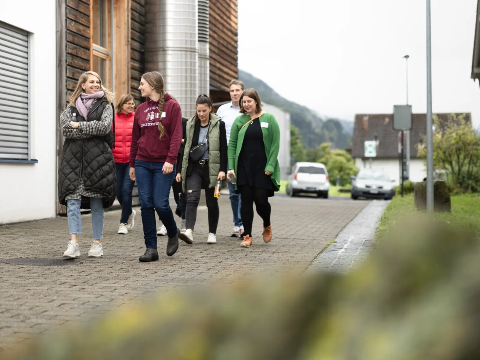 Eine Gruppe von Menschen geht auf einem gepflasterten Weg auf dem Campus der Universität Liechtenstein. Im Hintergrund sind Gebäude und eine bergige Landschaft zu sehen.