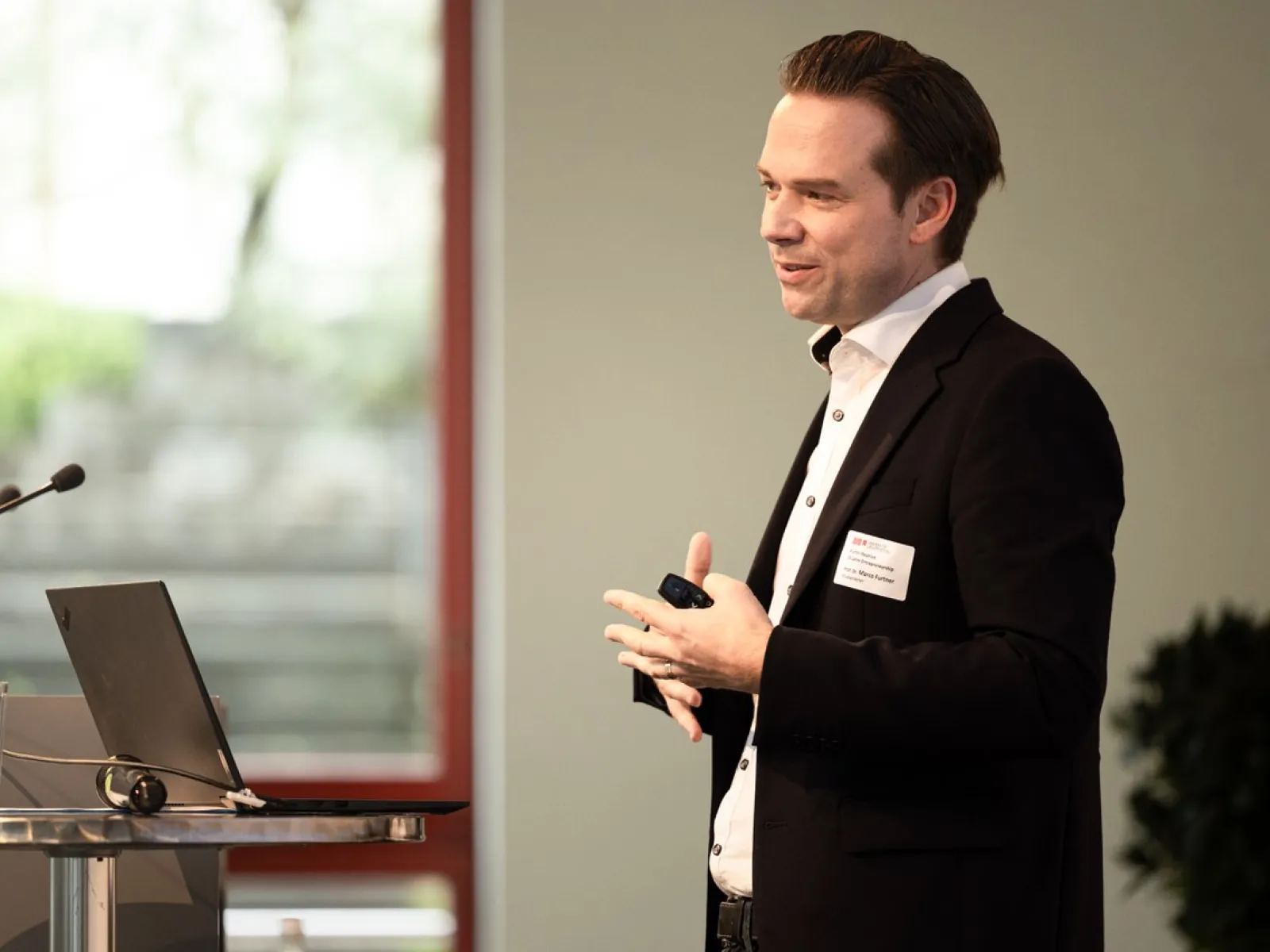 Ein Redner im Anzug hält einen Vortrag auf einer Veranstaltung an der Universität Liechtenstein. Im Hintergrund sind ein Fenster und Pflanzen zu sehen.