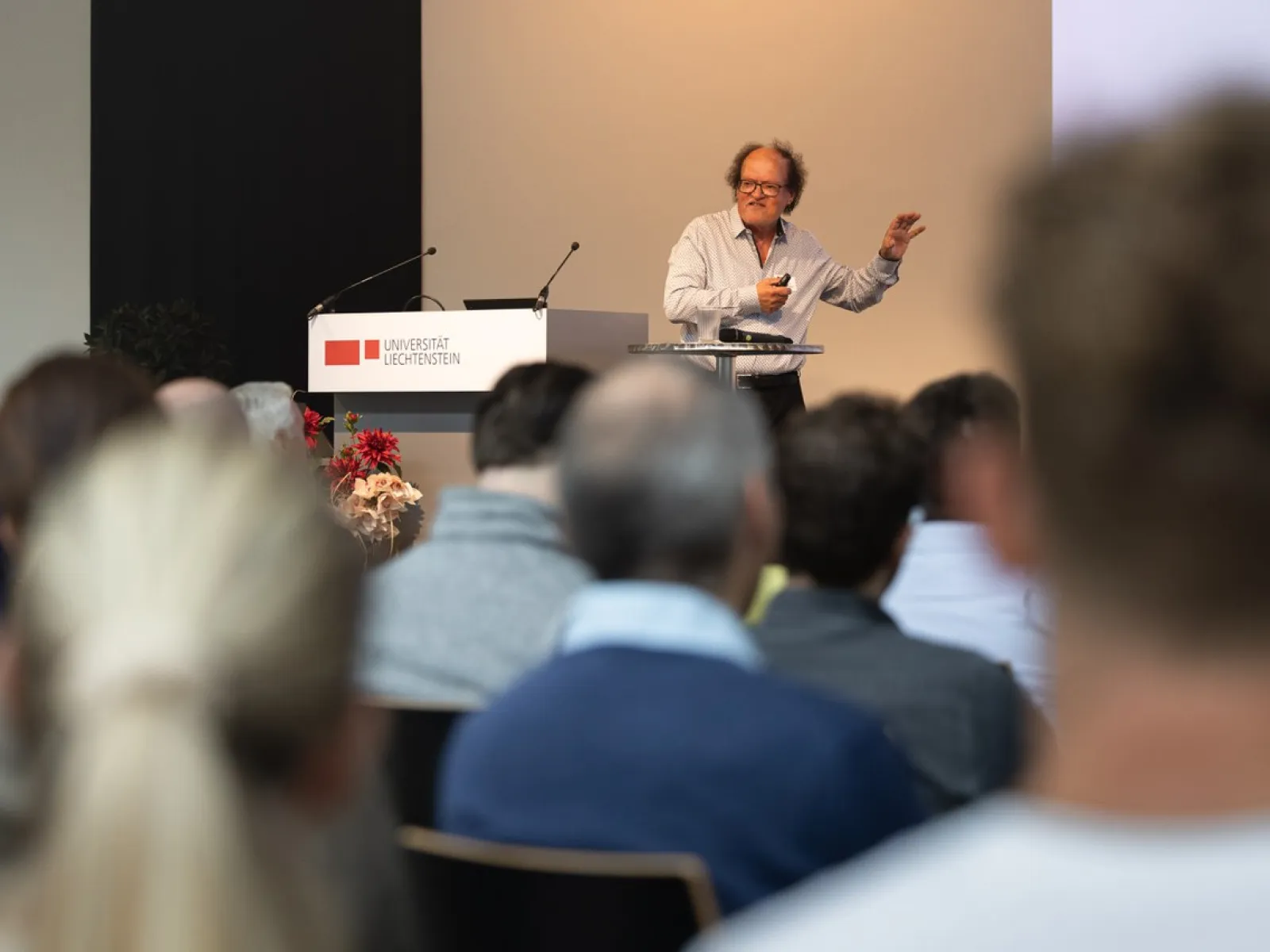 Ein Redner steht an einem Podium und spricht zu einem Publikum bei einer Veranstaltung an der Universität Liechtenstein. Im Hintergrund ist das Logo der Universität sichtbar.