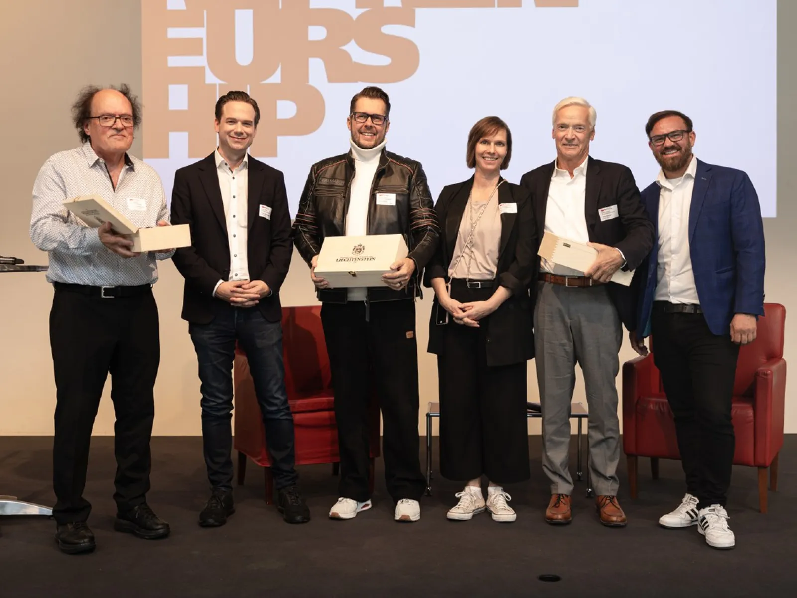 Sechs Personen stehen auf einer Bühne und halten Geschenke in den Händen. Im Hintergrund ist ein Bildschirm mit dem Wort "Entrepreneurship" zu sehen. Die Personen lächeln und tragen Namensschilder.