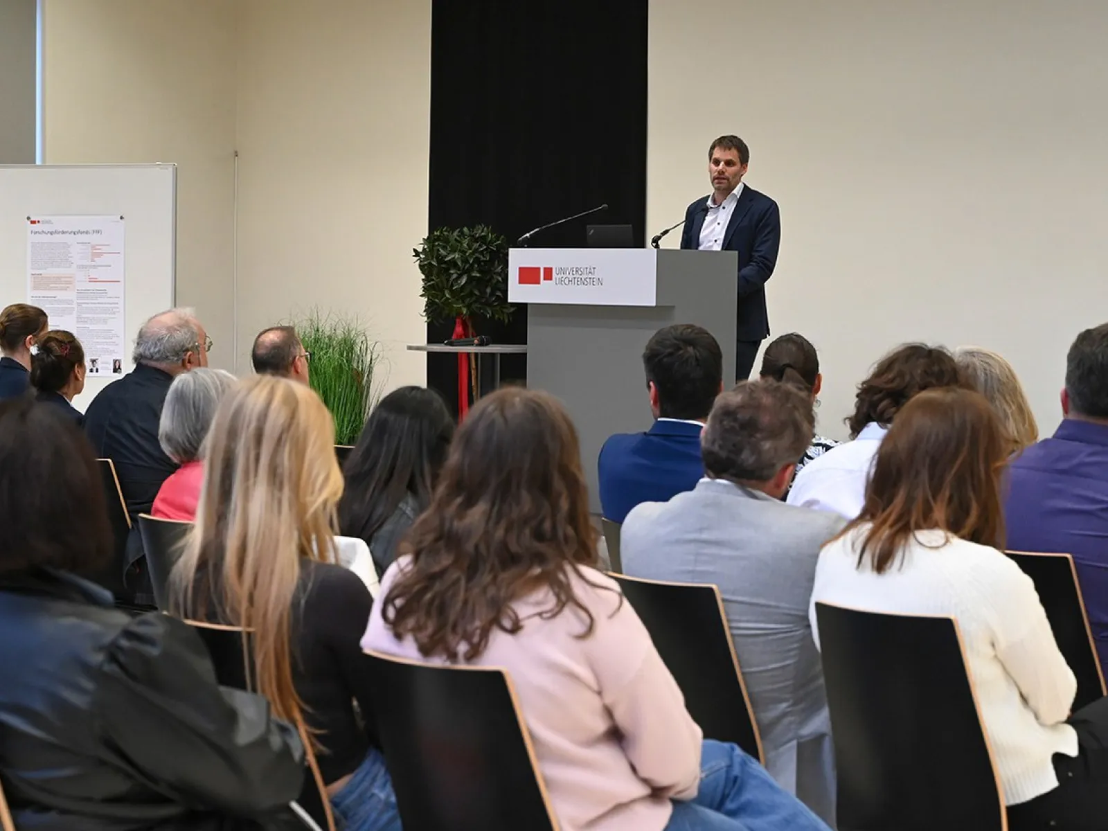 Rektor Christian Frommelt hält eine Rede am Rednerpult vor einem sitzenden Publikum in einem Konferenzraum. Im Hintergrund ist ein Poster sichtbar.