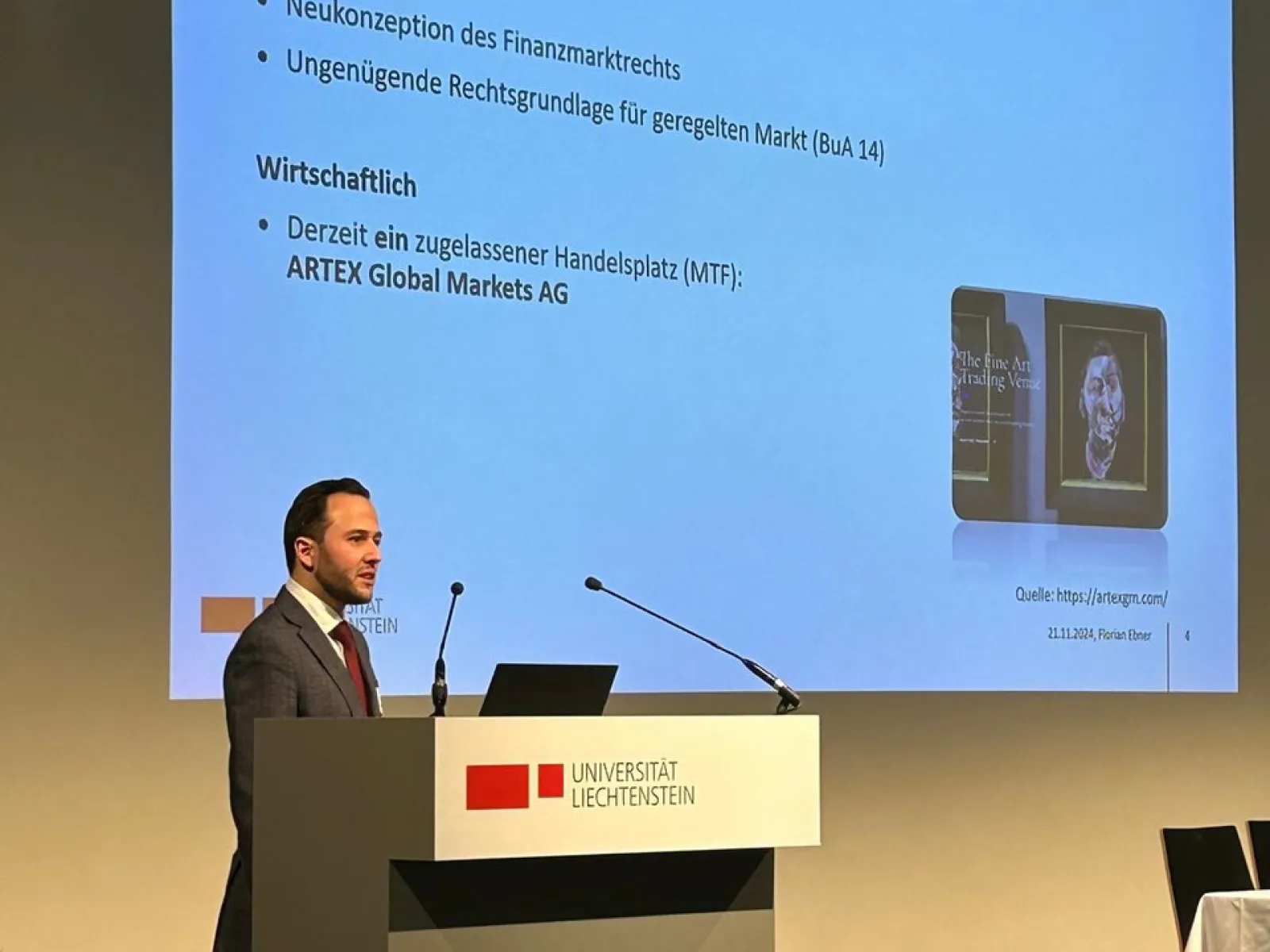 Ein Vortragender steht an einem Rednerpult der Universität Liechtenstein und präsentiert eine Folie mit dem Titel "Hintergrund". Die Folie enthält Informationen zur rechtlichen und wirtschaftlichen Neukonzeption des Finanzmarktrechts.