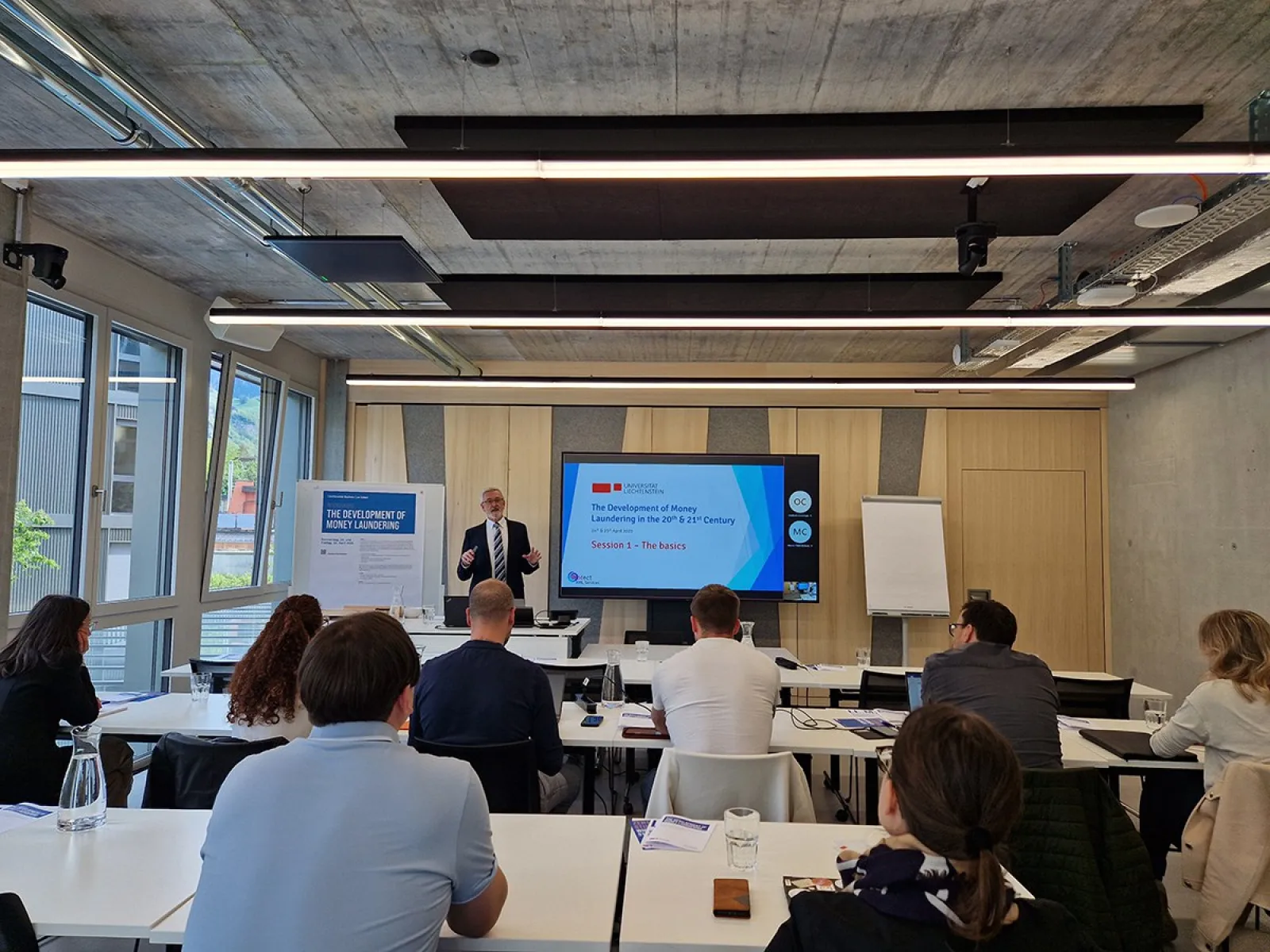 Ein Dozent hält einen Vortrag über Geldwäscherei im 20. und 21. Jahrhundert vor einer Gruppe von Teilnehmenden in einem modernen Seminarraum der Universität Liechtenstein. Im Hintergrund ist eine Präsentation auf einem Bildschirm zu sehen.