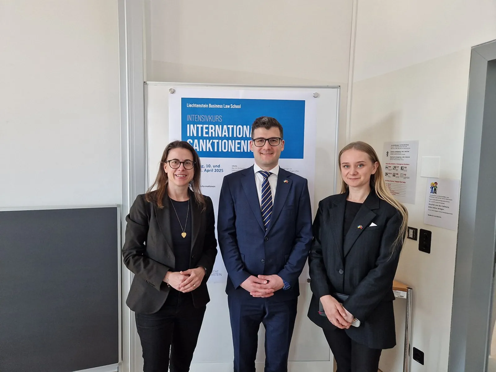 Drei Personen stehen vor einem Poster mit der Aufschrift "Intensivkurs Internationales Sanktionenrecht". Sie tragen formelle Kleidung und lächeln in die Kamera.