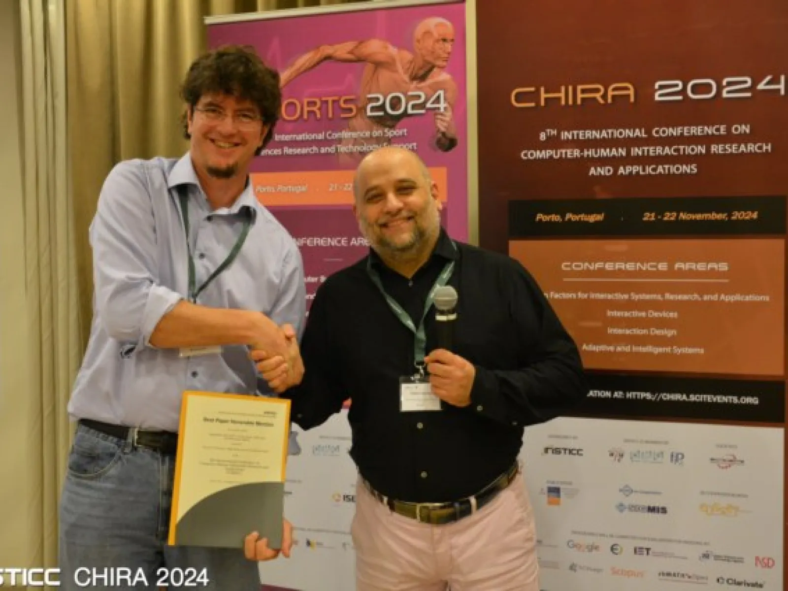 Zwei Männer stehen vor Bannern der CHIRA 2024 Konferenz. Einer hält ein Zertifikat, während der andere ein Mikrofon hält. Beide lächeln und schütteln sich die Hand. Im Hintergrund sind Details zur Konferenz in Porto, Portugal, sichtbar.